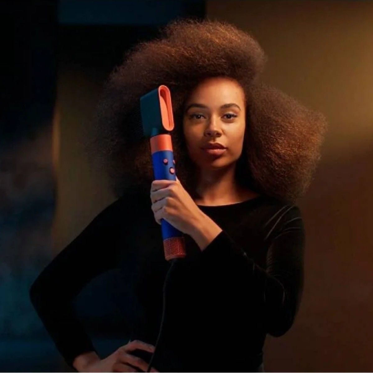 Donna con capelli voluminosi tiene in mano un asciugacapelli blu e arancione.