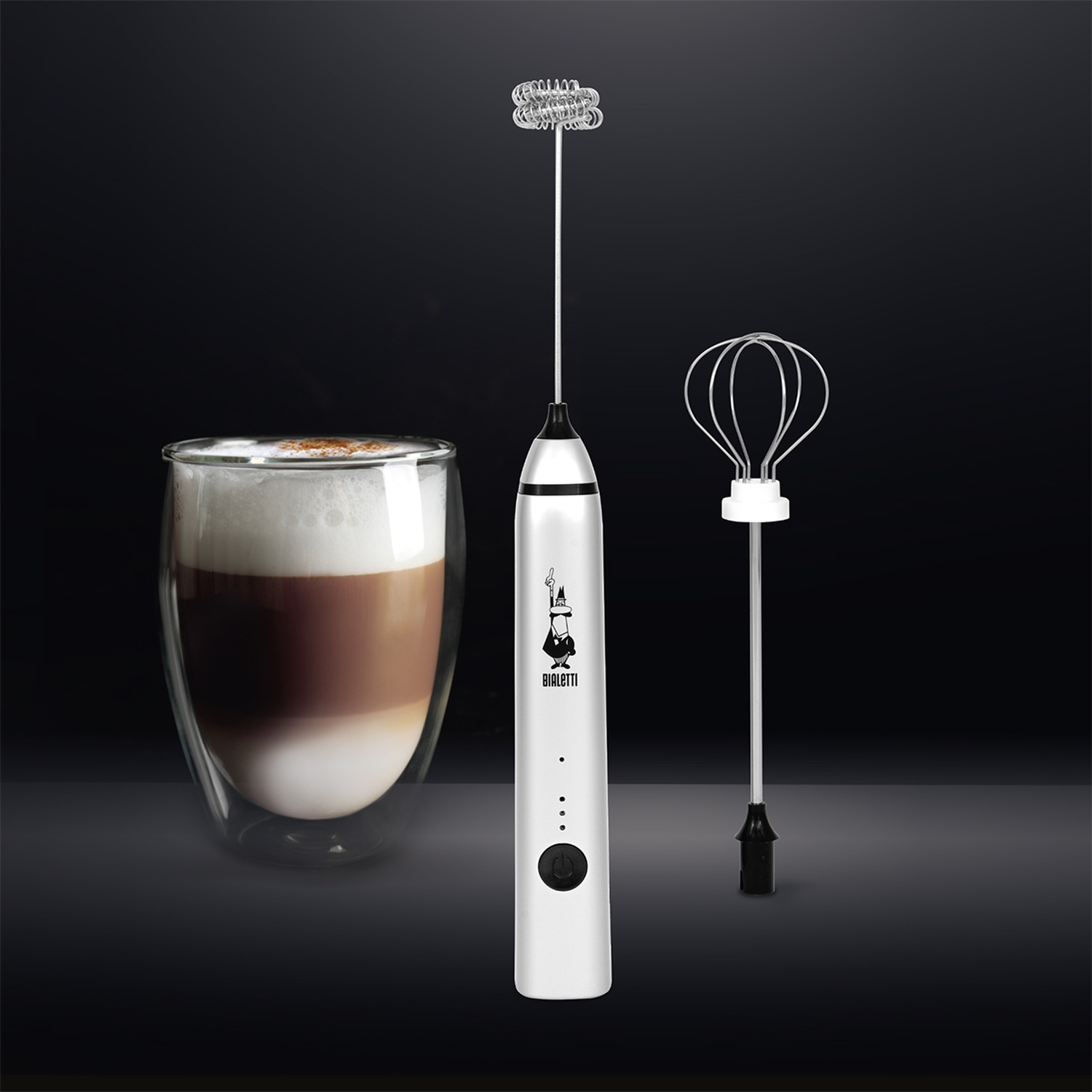 Een Bialetti melkopschuimer met twee opzetstukken naast een glas gelaagde latte.