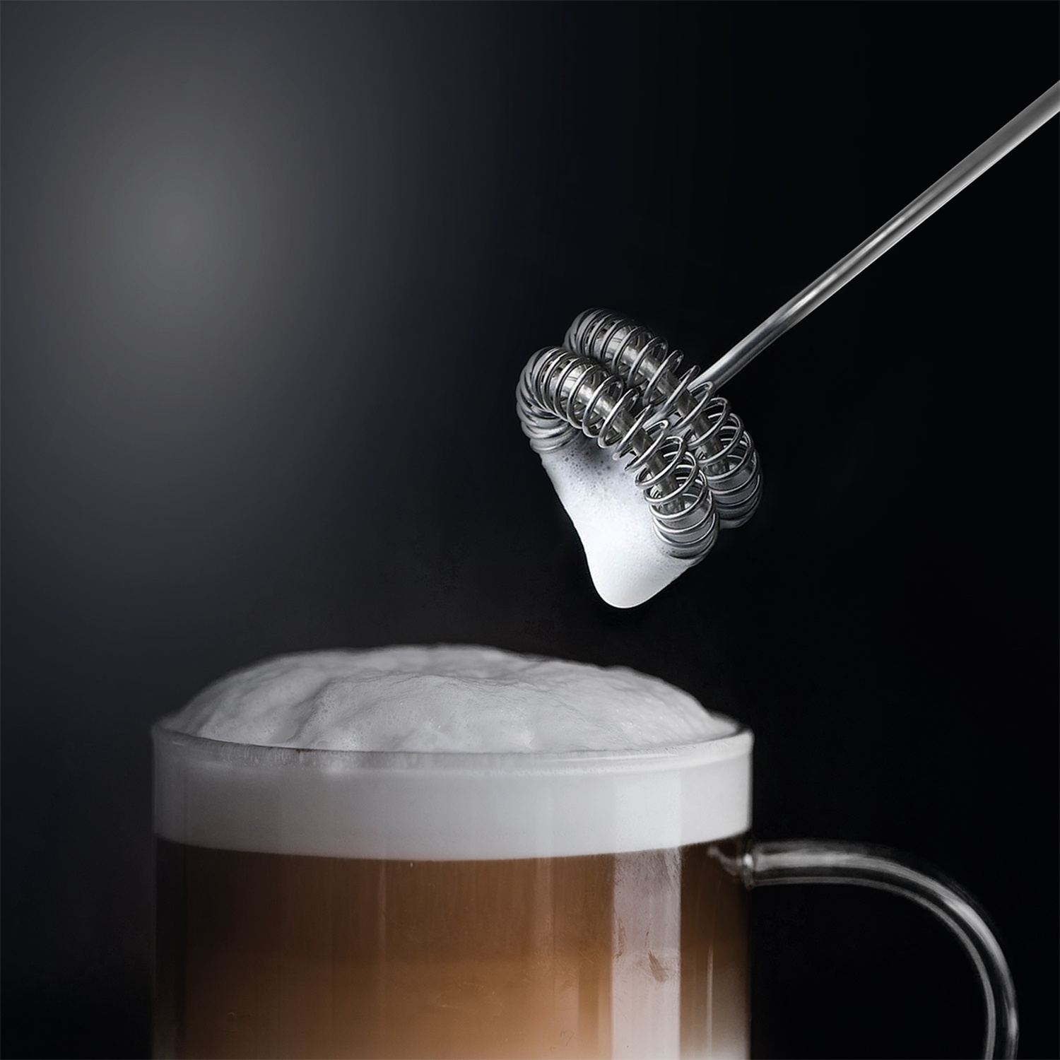 Een opschuimer creëert schuim op een latte in een doorzichtige mok tegen een donkere achtergrond.