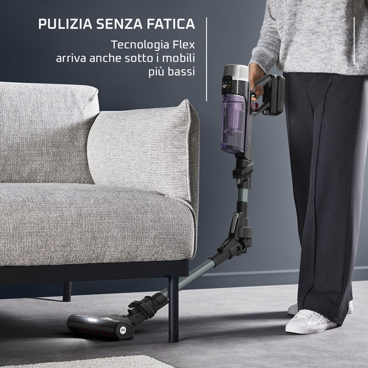 Una persona usa una aspiradora inalámbrica para limpiar debajo de un sofá gris.