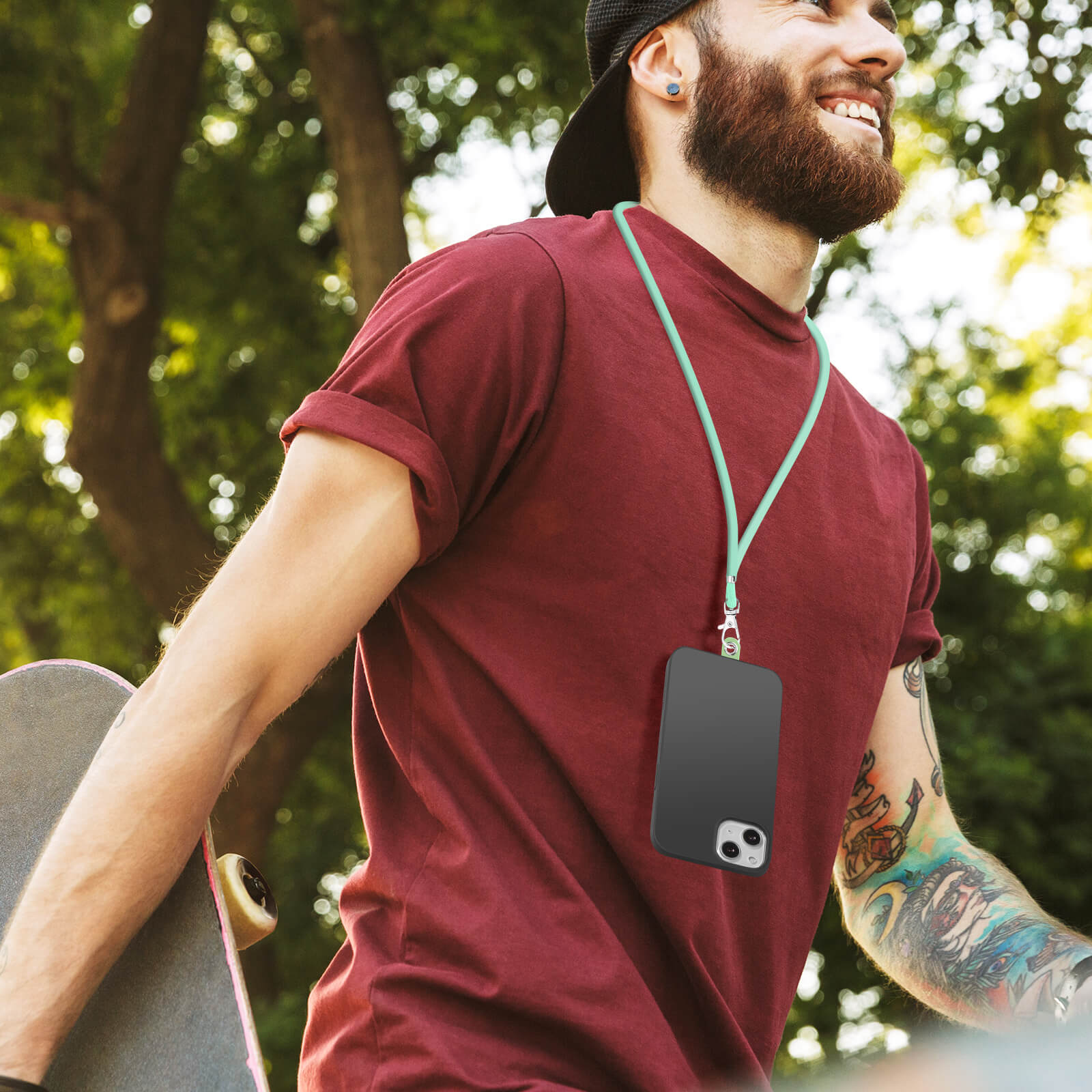 Un uomo con t-shirt bordeaux e cappellino sorride, porta il telefono con un laccetto. Tiene uno skateboard.