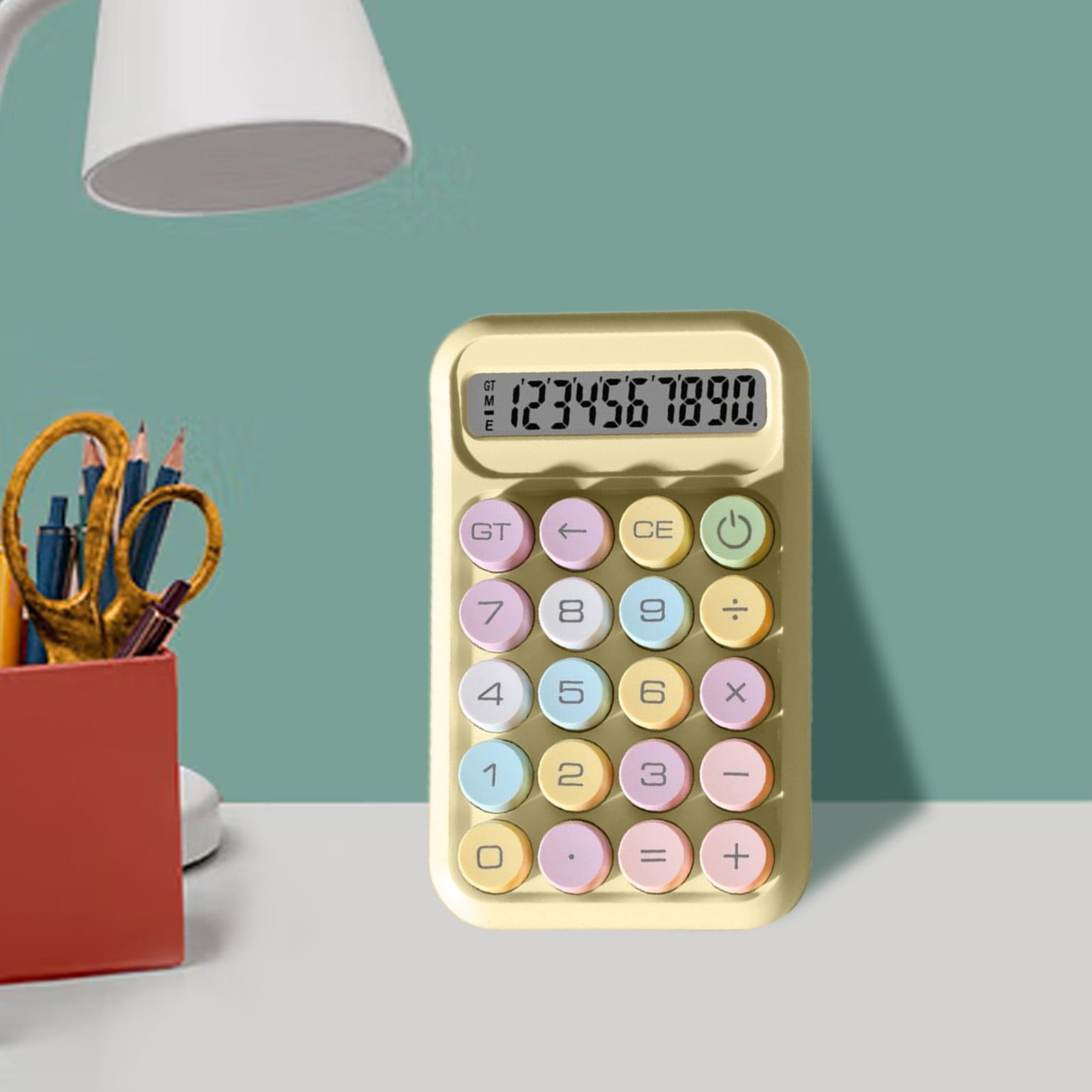 Ein pastellgelber Taschenrechner mit bunten Tasten steht auf einem weißen Schreibtisch. Eine Lampe und ein roter Halter mit Schere und Stiften sind in der Nähe.