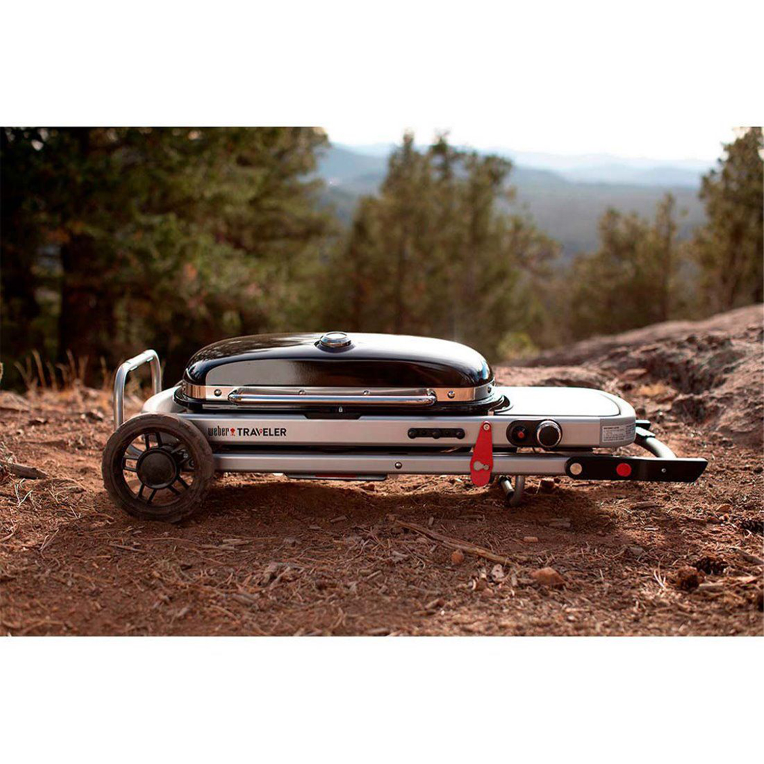 Srebrny grill Weber Traveler stoi na suchej ziemi, z drzewami i wzgórzami w tle.