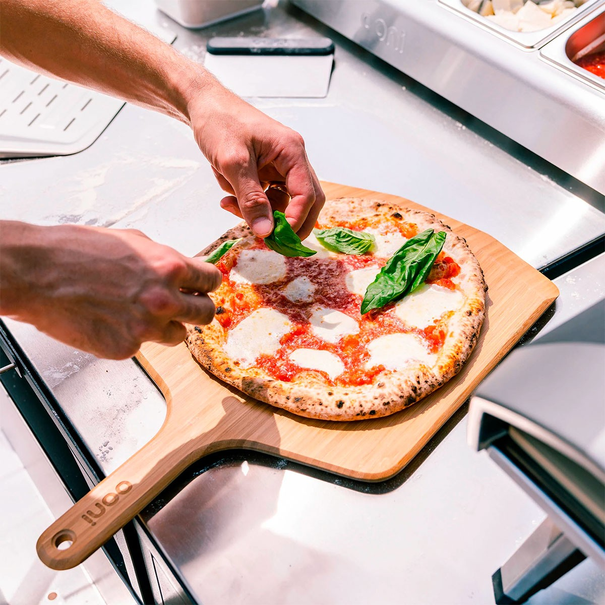 Mani che mettono basilico su una pizza appena sfornata su una pala di legno, pronta per essere servita.