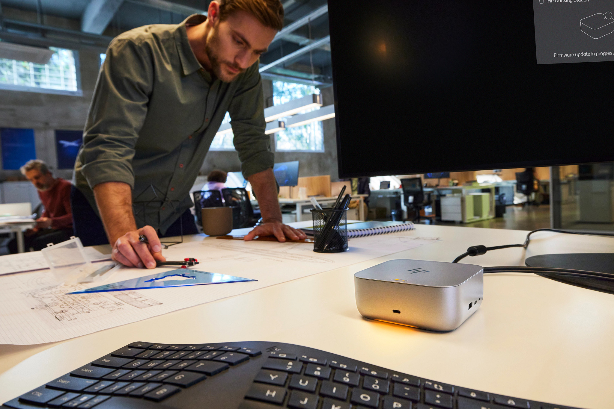 Mann in grünem Hemd prüft Baupläne am Schreibtisch mit Dockingstation und Tastatur.