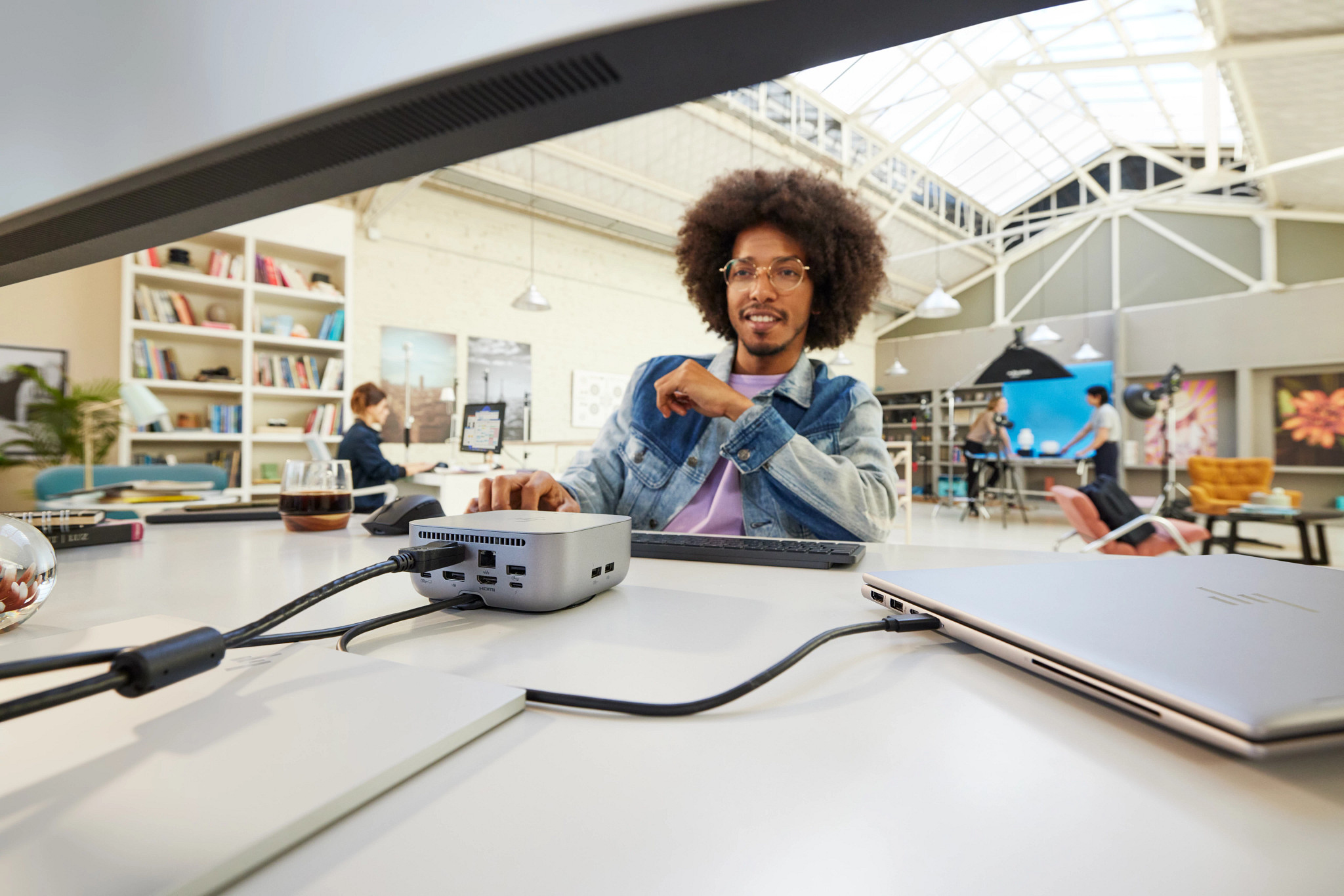 Mann mit Afro lächelt in die Kamera, Schreibtisch mit Computer, Kabeln und Laptop.