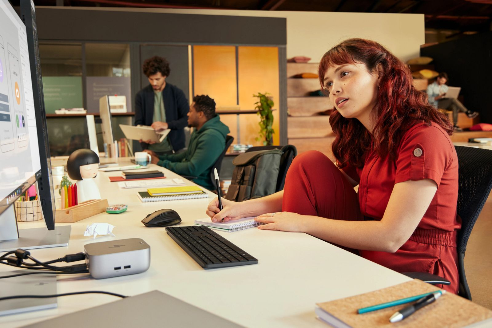 Menschen arbeiten an Schreibtischen in einem modernen Büro. Eine Frau in Rot schreibt in ein Notizbuch.
