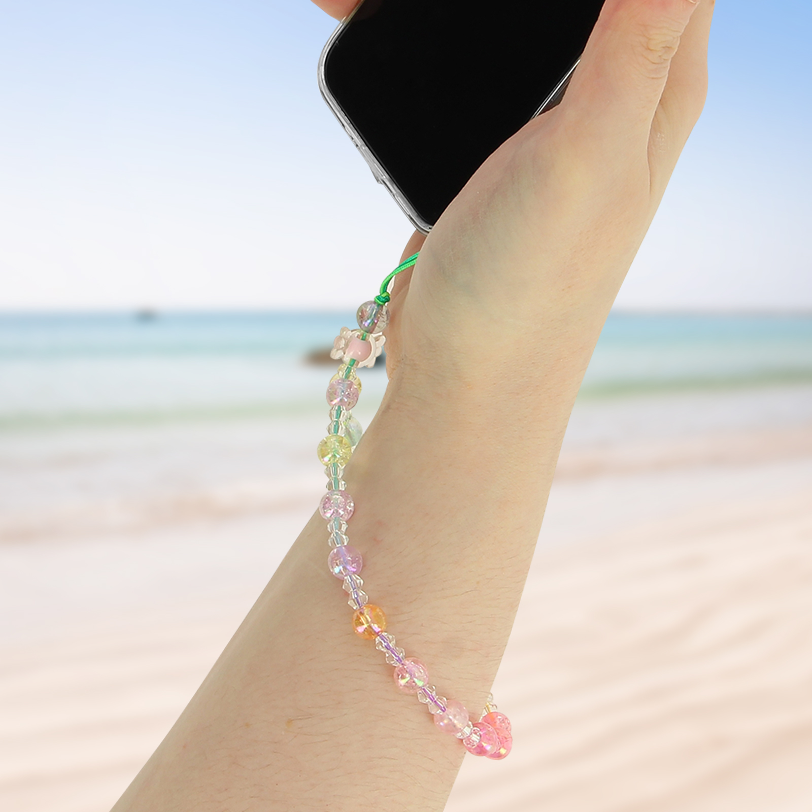 Mano che tiene il telefono con cinturino di perline colorate e ciondolo a farfalla, sfondo spiaggia.