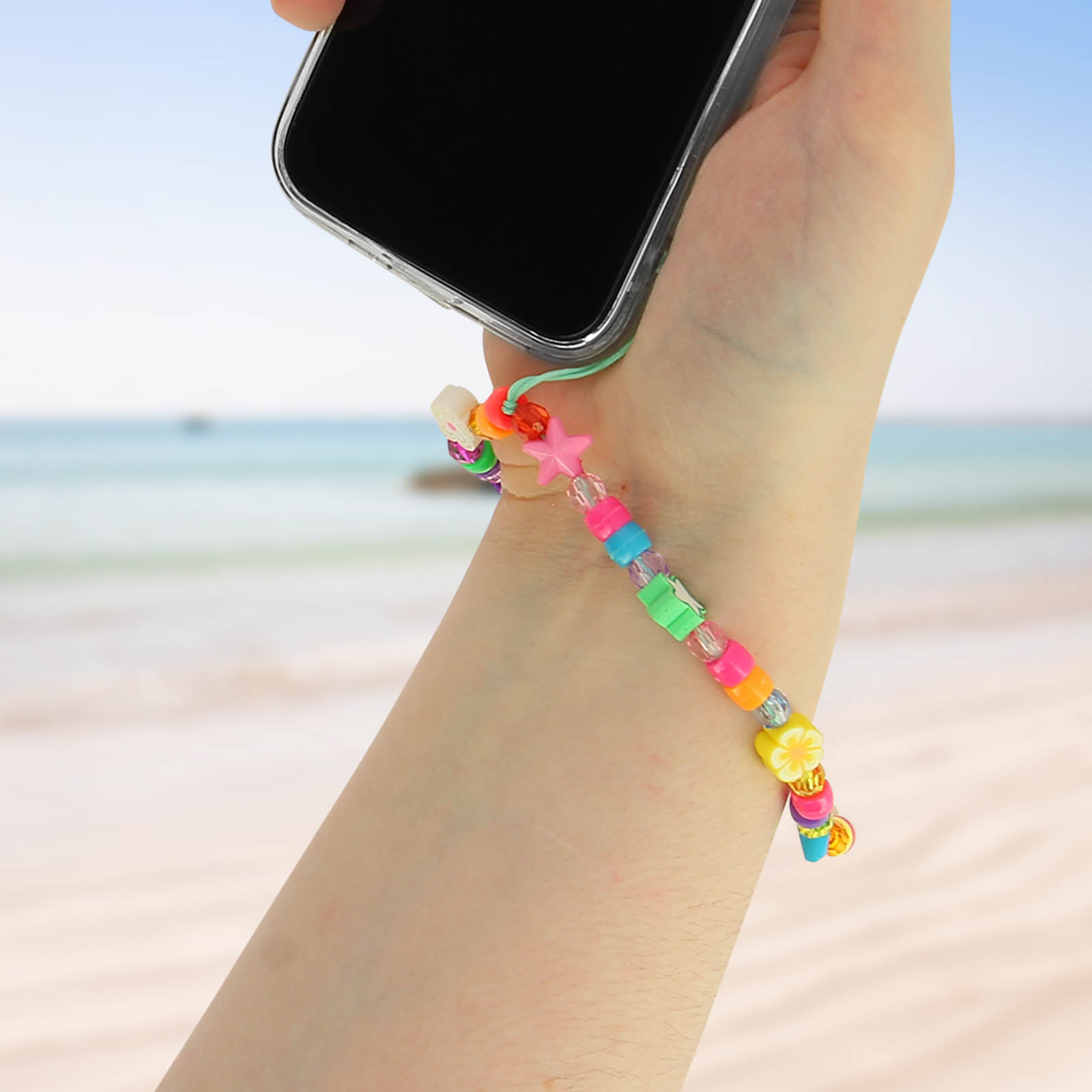 Una mano tiene un telefono con un ciondolo di perline colorate su sfondo spiaggia.