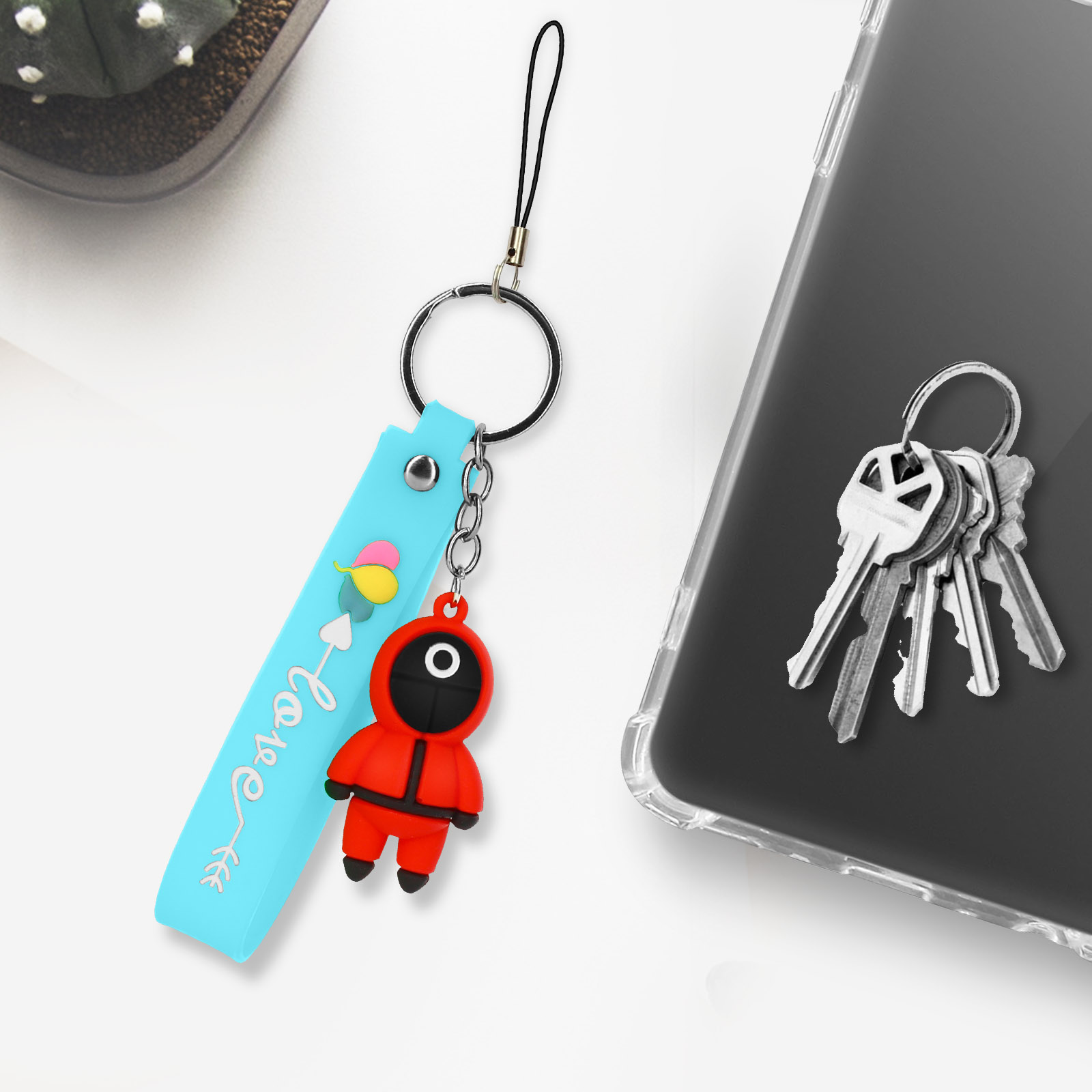 Un portachiavi blu con un ciondolo personaggio di Squid Game rosso pende accanto a un telefono con chiavi.