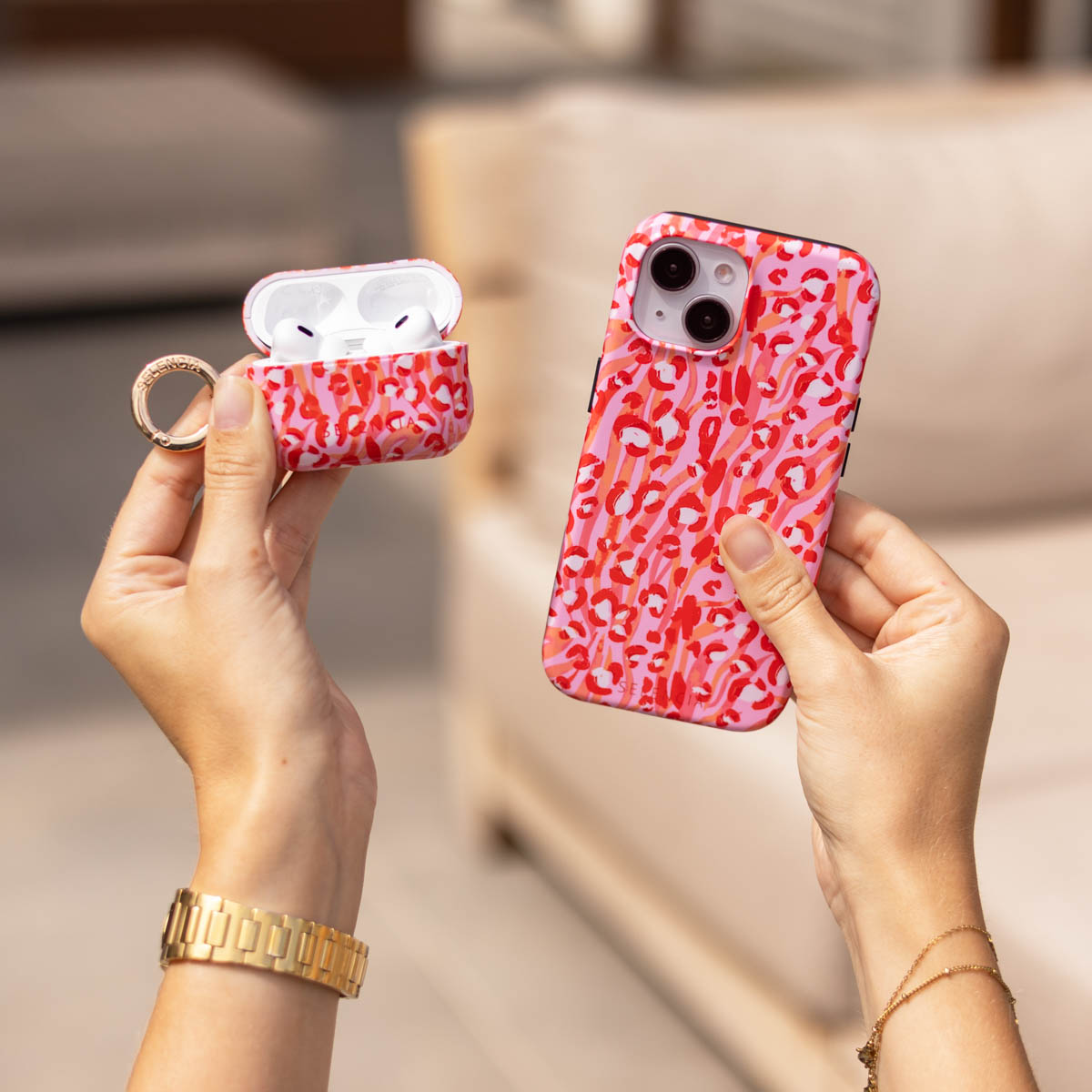 Le mani tengono una custodia per telefono rosa con stampa leopardata e una custodia per AirPods con anello dorato.