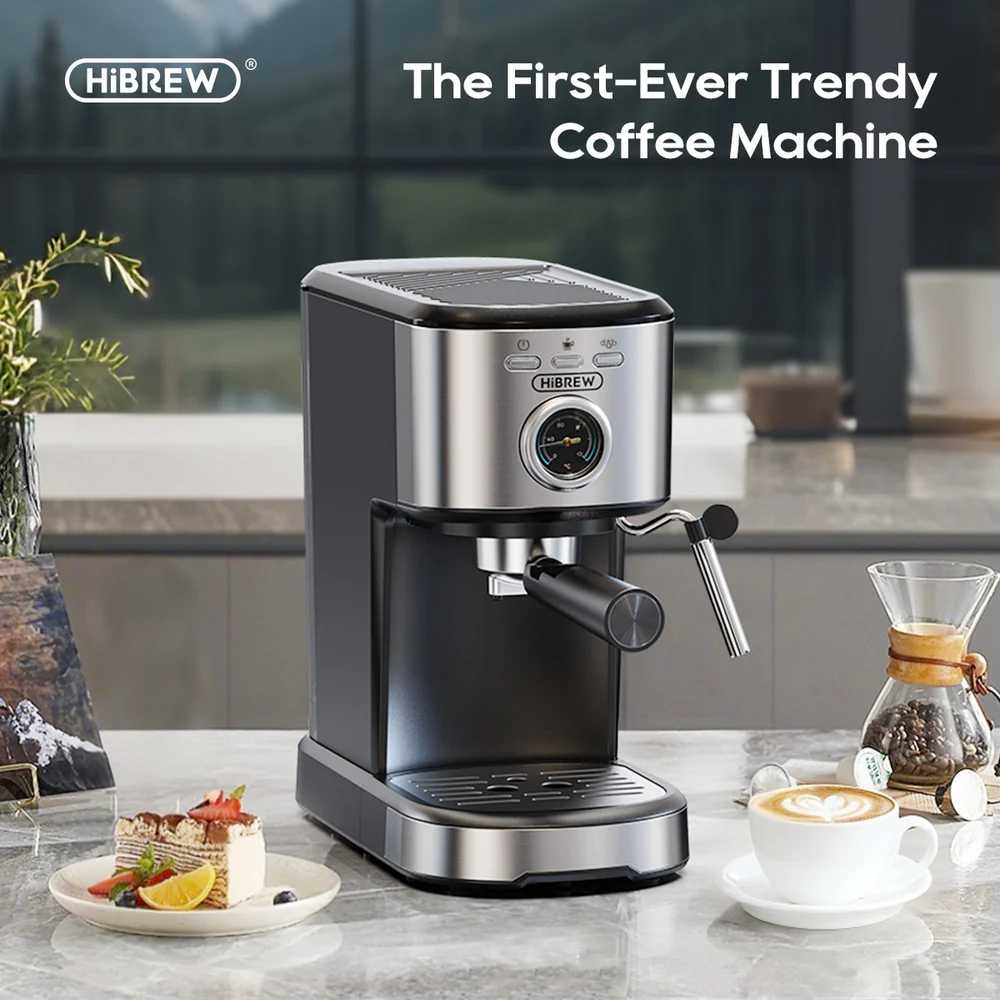 Una cafetera HIBREW negra y plateada está sobre un mostrador con un trozo de pastel y un café con leche.