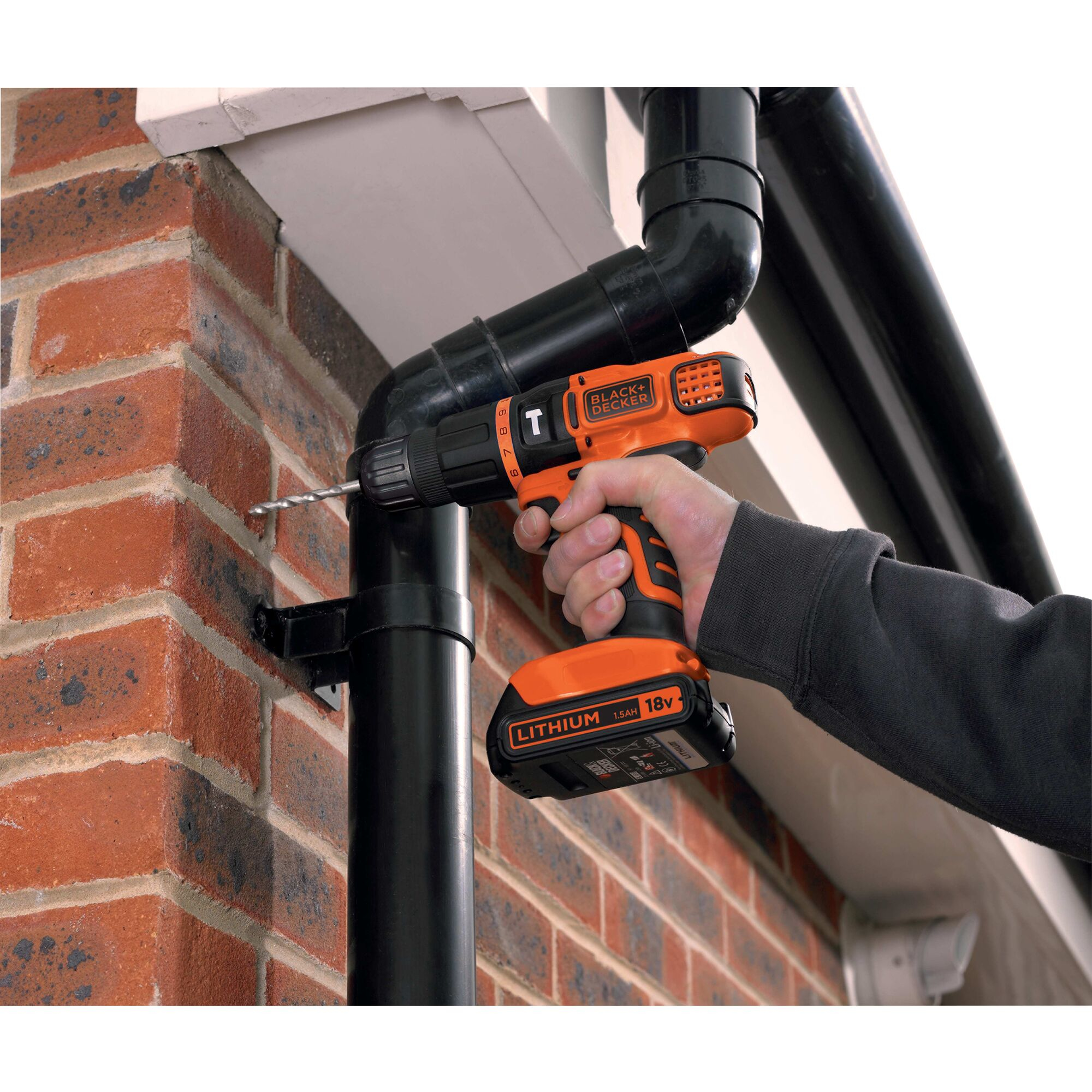 Un trapano Black+Decker fora un tubo nero contro un muro di mattoni.