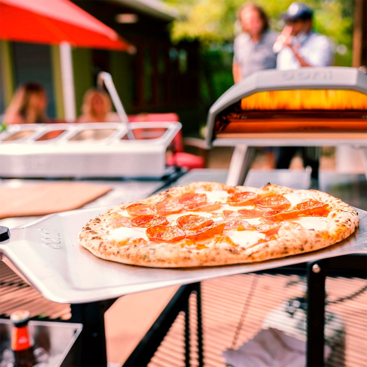 Eine Peperoni-Pizza liegt auf einer Metallschaufel vor einem Pizzaofen im Freien mit verschwommenen Personen im Hintergrund.