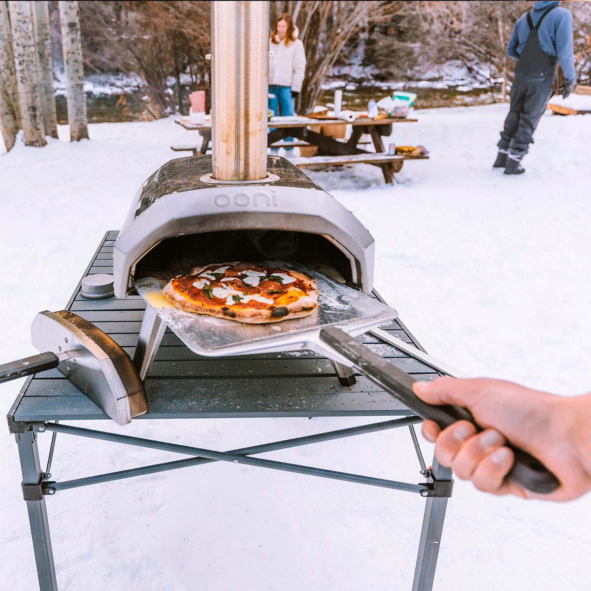 Eine Pizza wird von einer Person mit einem Pizzaschieber aus einem Ooni-Ofen im Schnee genommen.