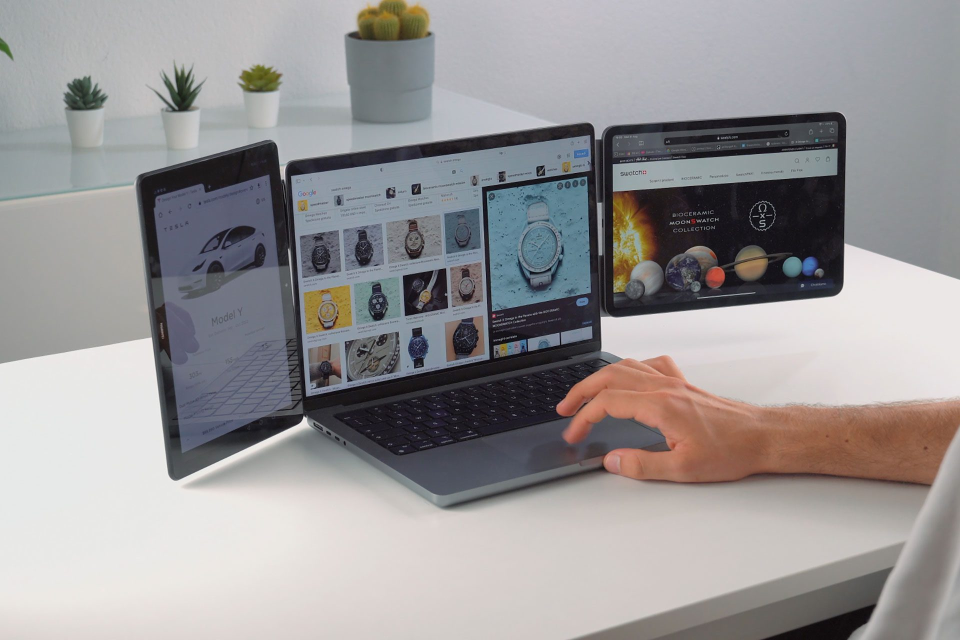 Una persona usa un laptop con due tablet laterali che mostrano siti web di Tesla, orologi e pianeti.