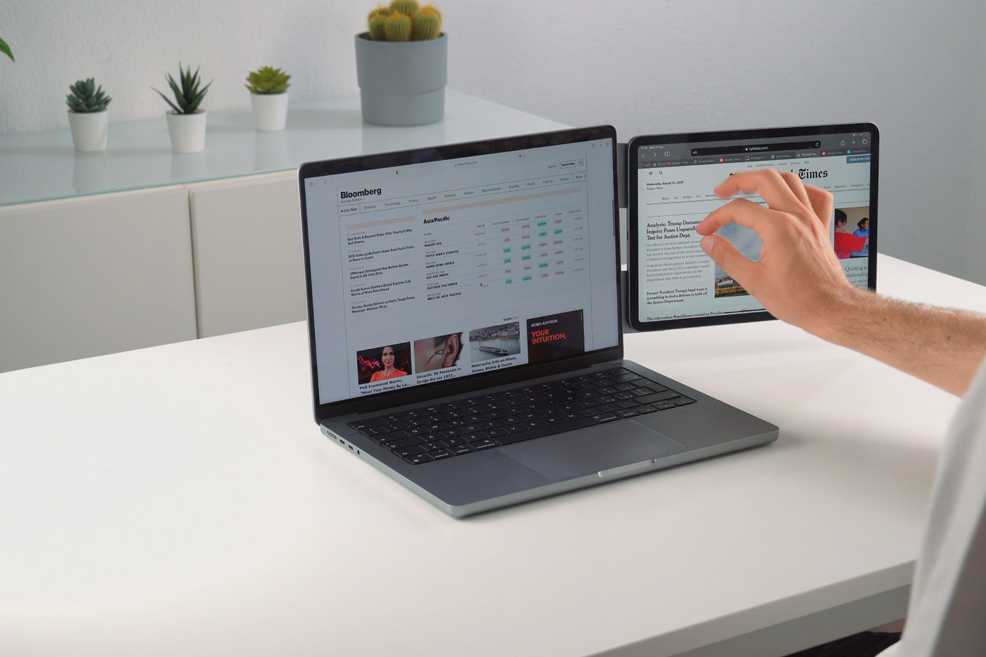 Una mano interagisce con un tablet che mostra il New York Times accanto a un laptop che mostra Bloomberg.