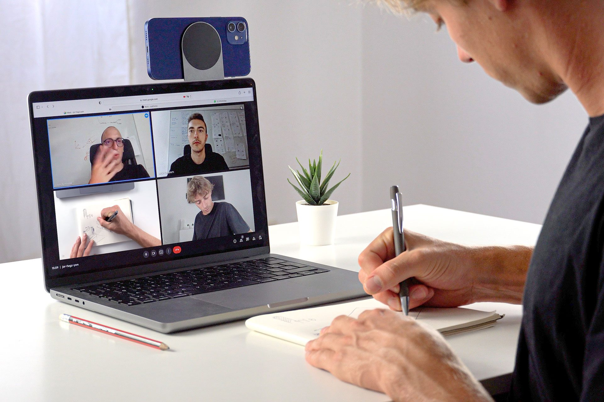 Uomo in videochiamata, prende appunti. Quattro persone visibili sullo schermo del laptop.