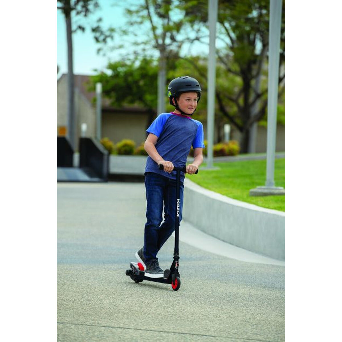 Ragazzo con casco guida monopattino nero su sentiero pavimentato con erba e alberi.