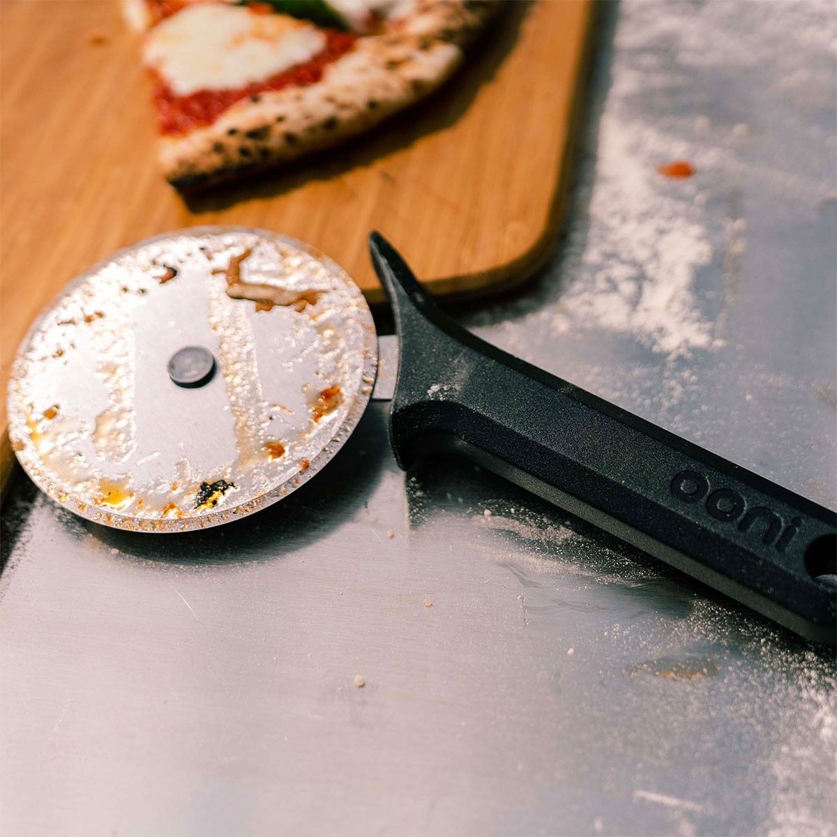 Ein Pizzaschneider mit schwarzem Griff liegt auf einer Metalloberfläche neben einem Pizzastück.