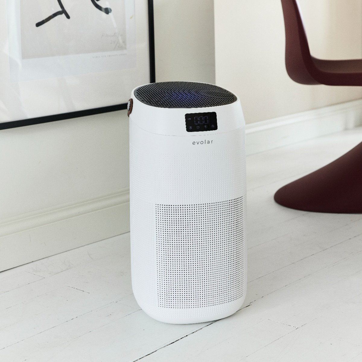 A white air purifier sits on a wooden floor next to artwork and a chair.