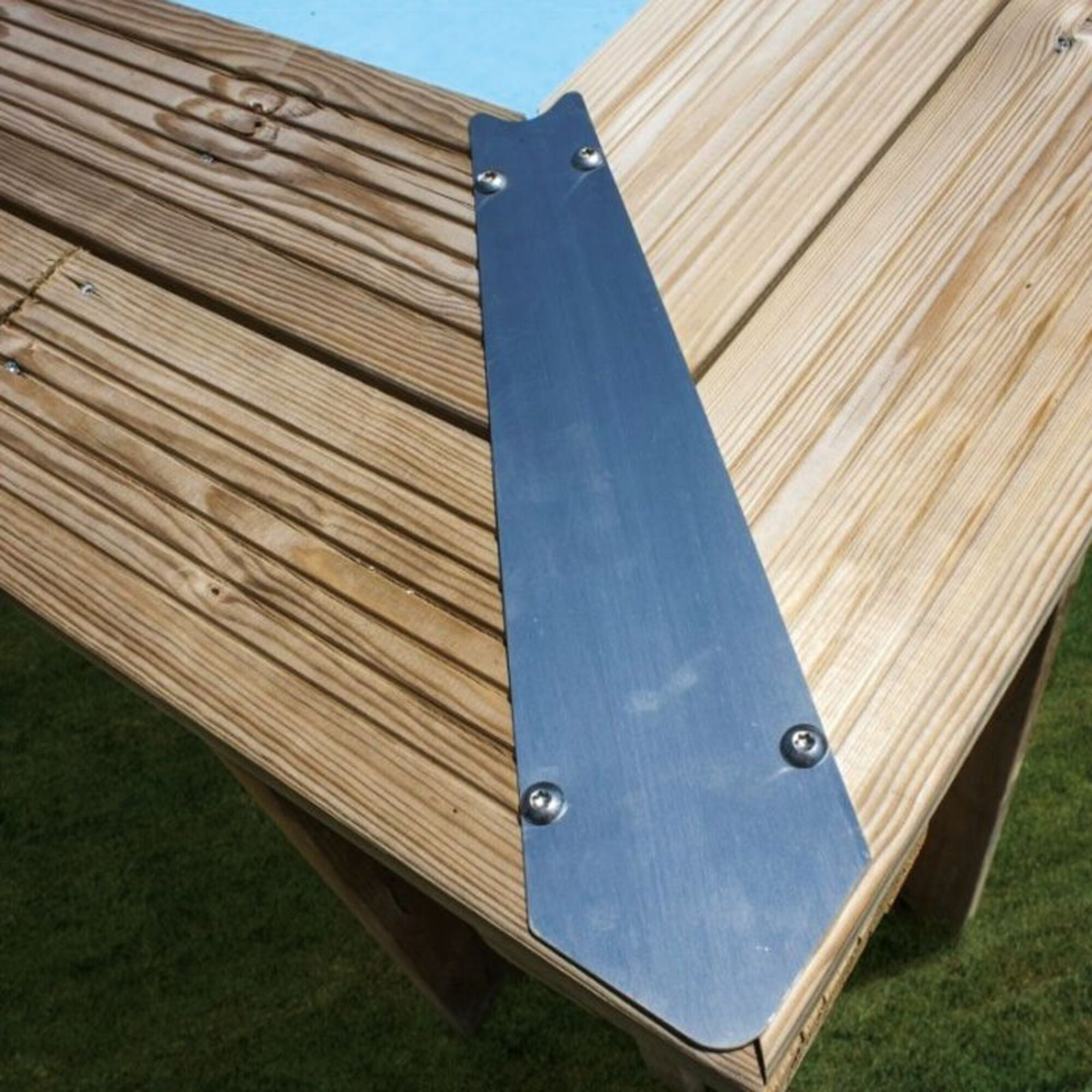 Una placa metálica refuerza la esquina de una cubierta de madera. Césped verde debajo.