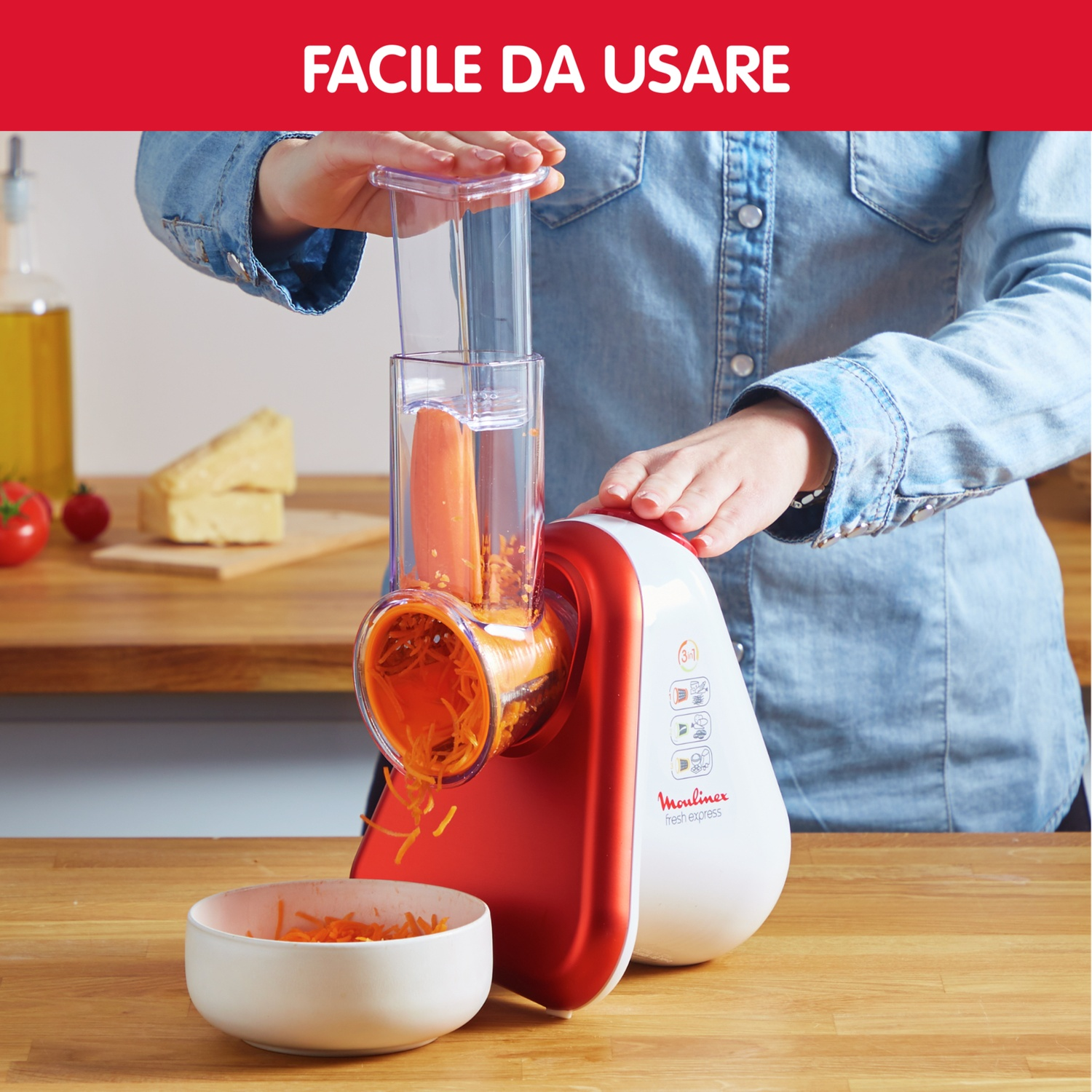 Un robot da cucina rosso e bianco grattugia una carota in una ciotola. Formaggio e pomodori vicini.