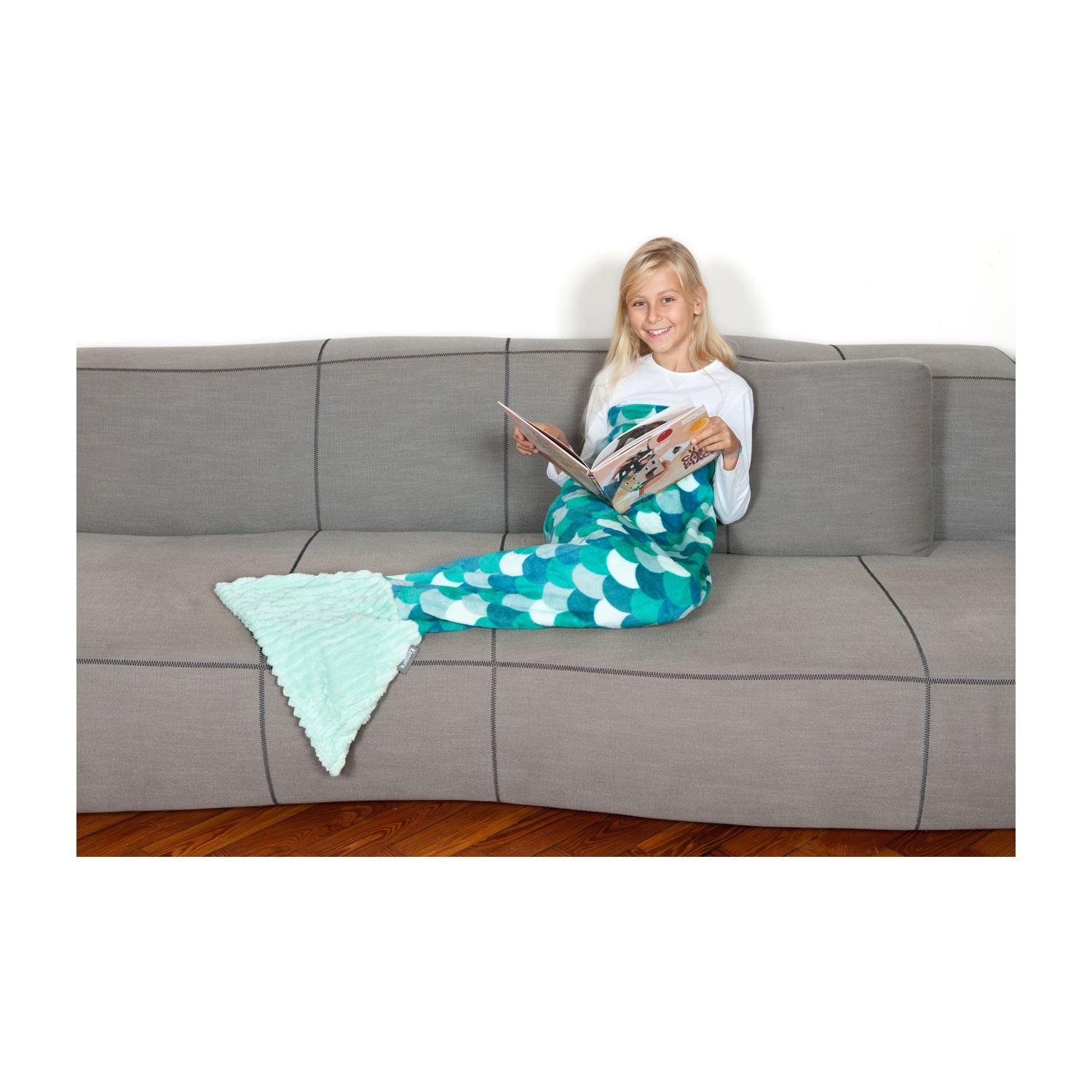 Una bambina seduta su un divano grigio legge un libro, avvolta in una coperta a coda di sirena.