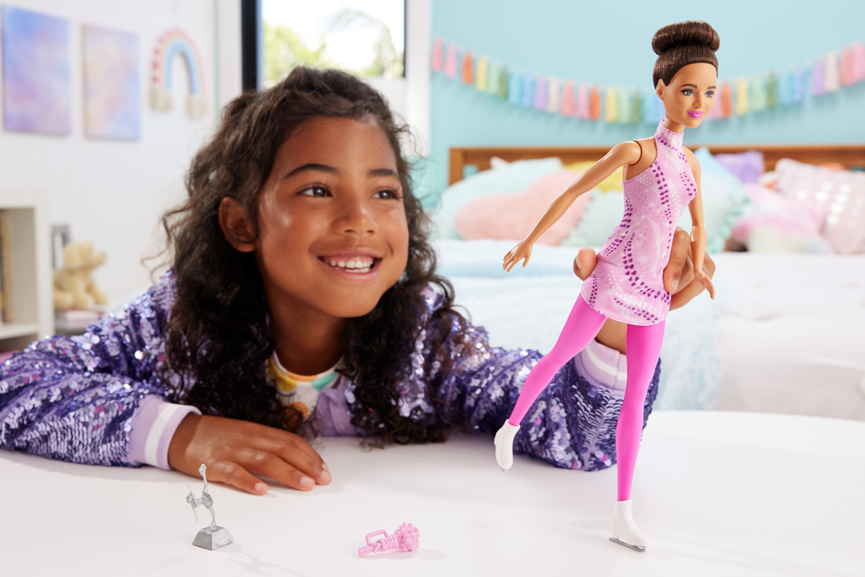 Una bambina con giacca scintillante sorride, guardando una bambola Barbie pattinatrice.
