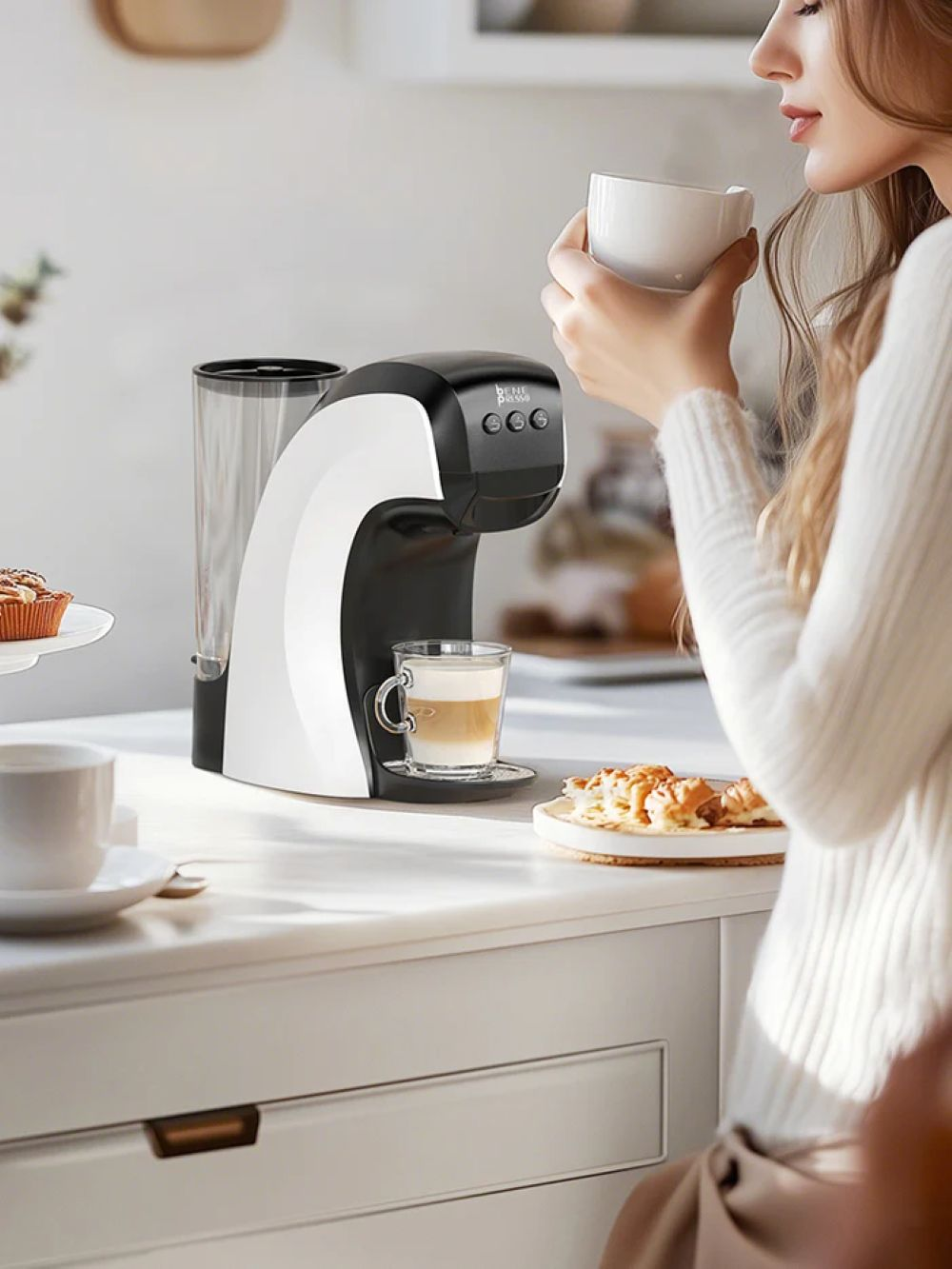 Una mujer disfruta de un café junto a una cafetera blanca y negra en una encimera de cocina.