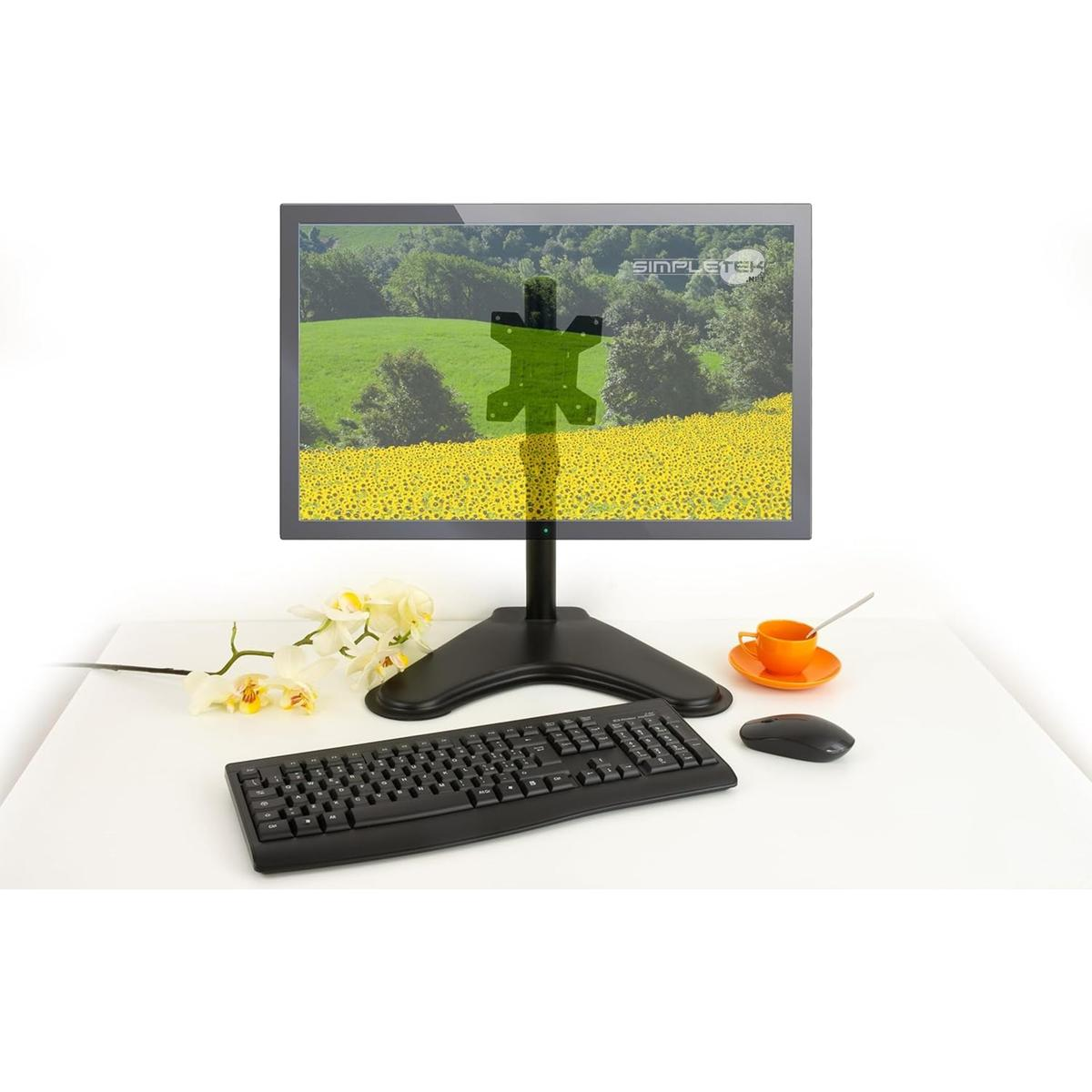 Un monitor per computer su un supporto mostra un campo di girasoli. Tastiera, mouse e fiore sono su una scrivania bianca.