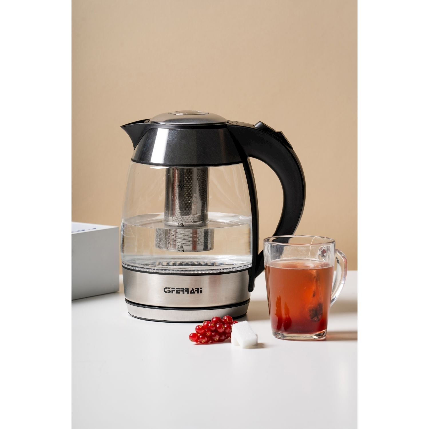 Szklany czajnik z wodą, kubek czerwonej herbaty, czerwone porzeczki i kostki cukru na białym stole.