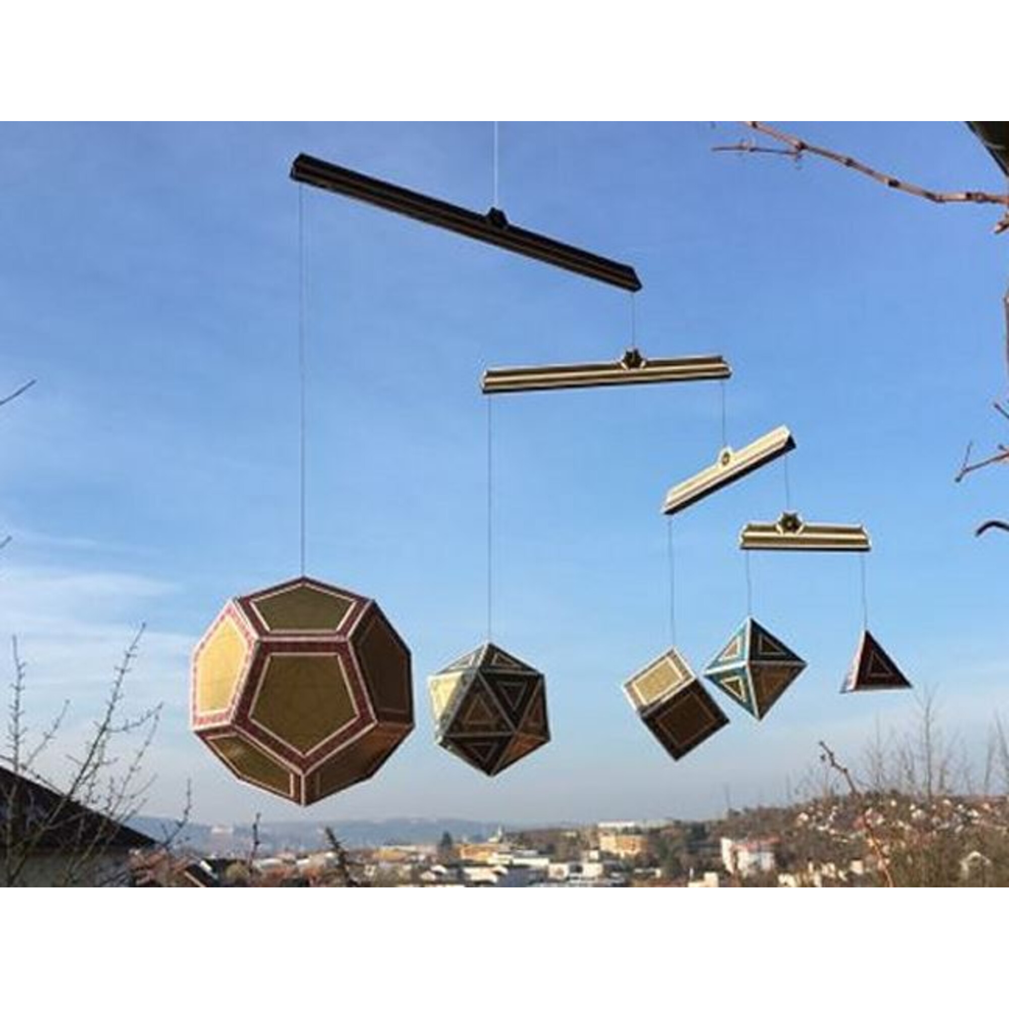 Geometrische Mobiles hängen vor blauem Himmel, mit einer Stadt im Hintergrund.