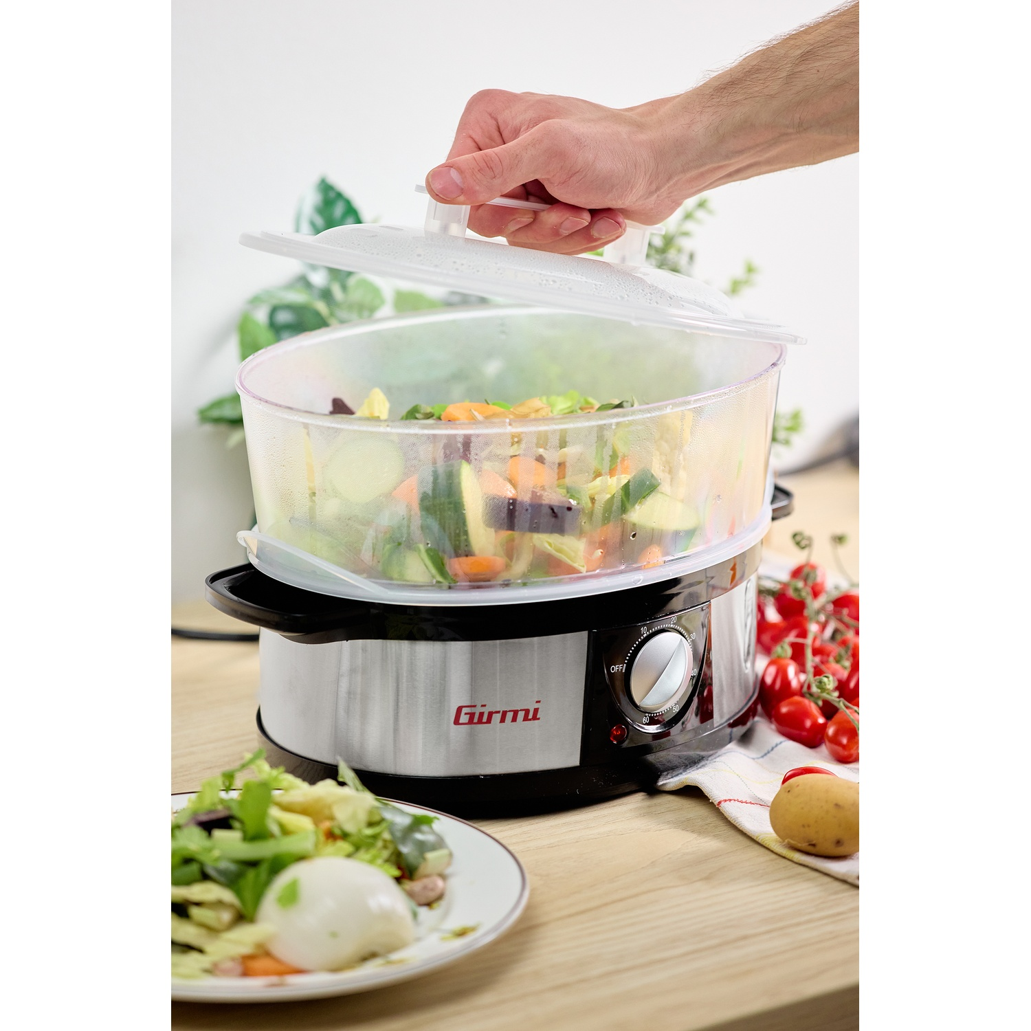 Una mano solleva il coperchio di un cuociriso pieno di verdure tritate. Un piatto di insalata è vicino.