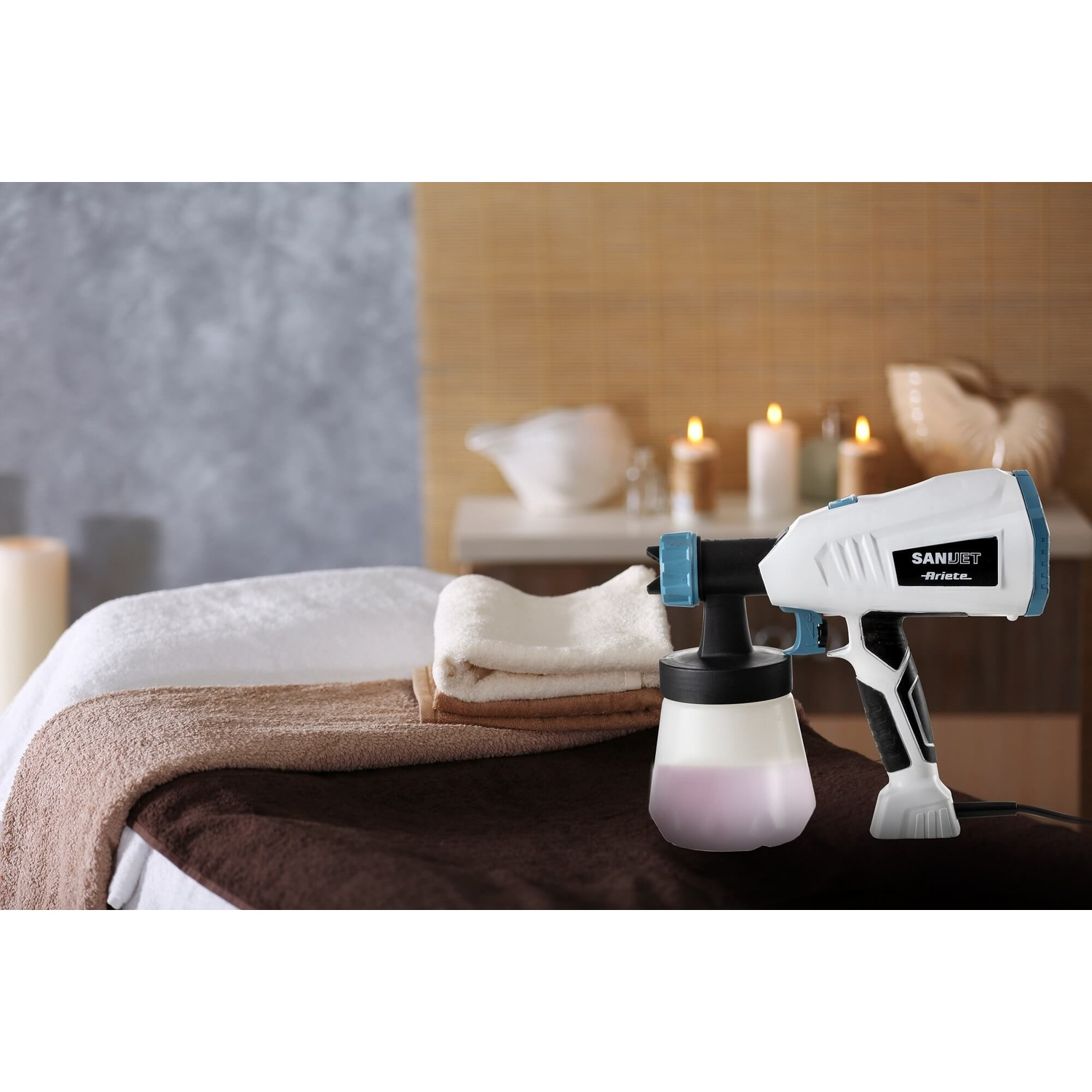 Pistola a spruzzo bianca e blu con liquido nel contenitore su asciugamani da spa, candele sullo sfondo.