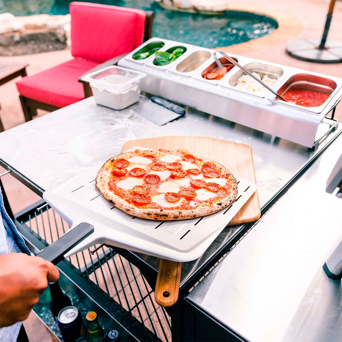 Una pizza al salame piccante su una pala, con condimenti in un vassoio. Piscina visibile.