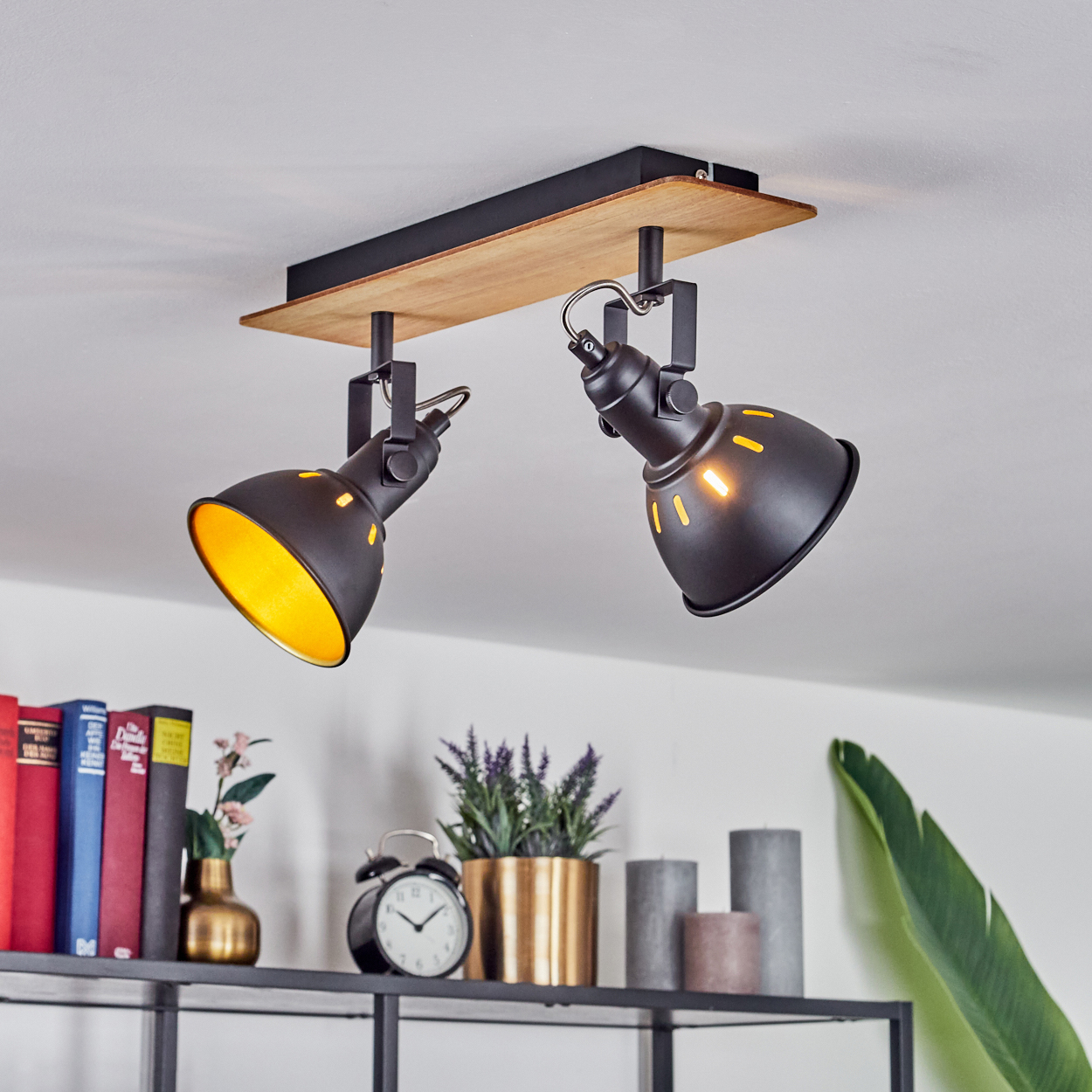 Ein schwarzer Doppelscheinwerfer mit Holzsockel ist an einer weißen Decke montiert. Darunter befinden sich Regale mit Büchern, einer Vase, einer Pflanze, Kerzen und einem Wecker.