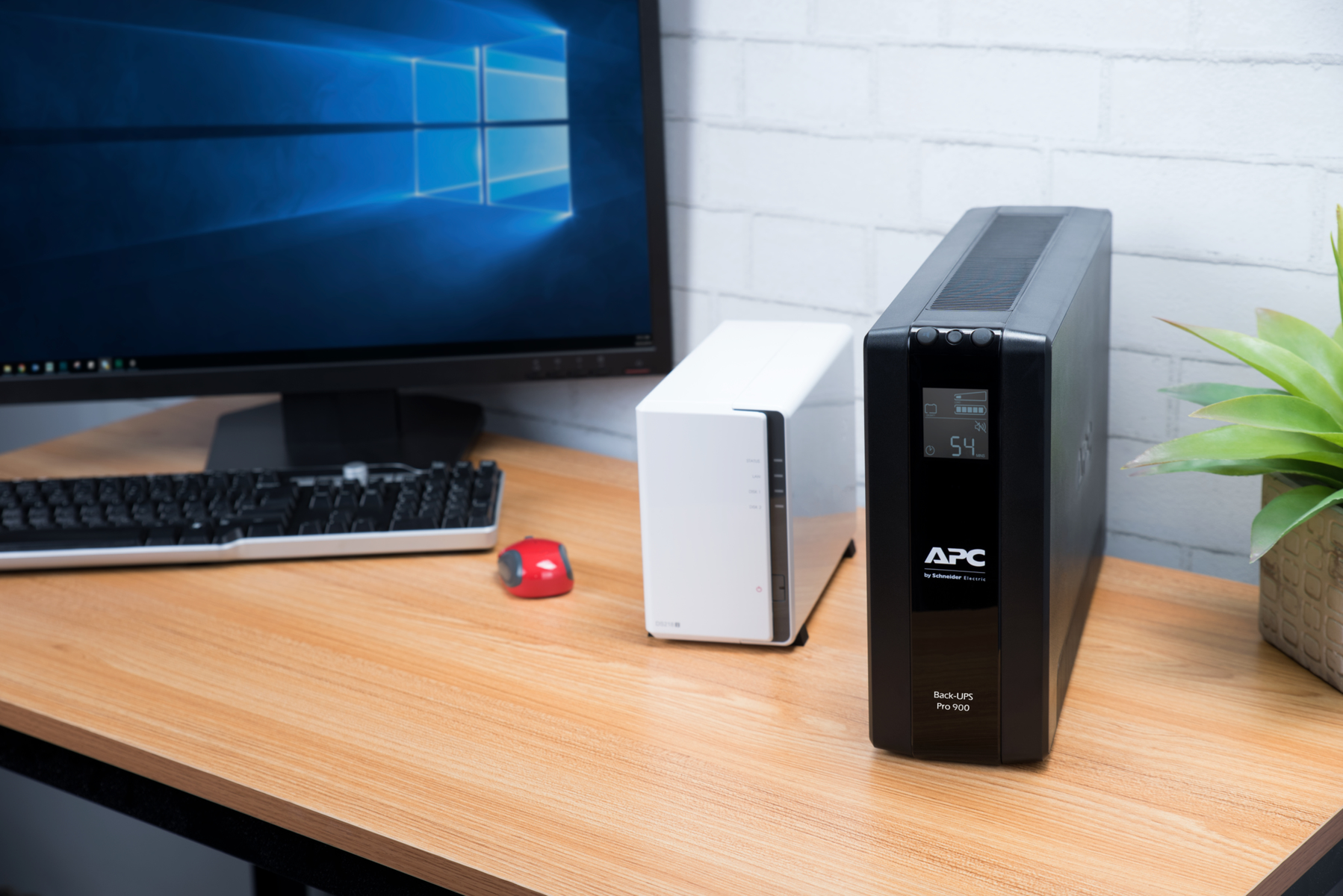 Ein Computermonitor, Tastatur, rote Maus, weißes NAS und schwarzer USV stehen auf einem Holztisch.