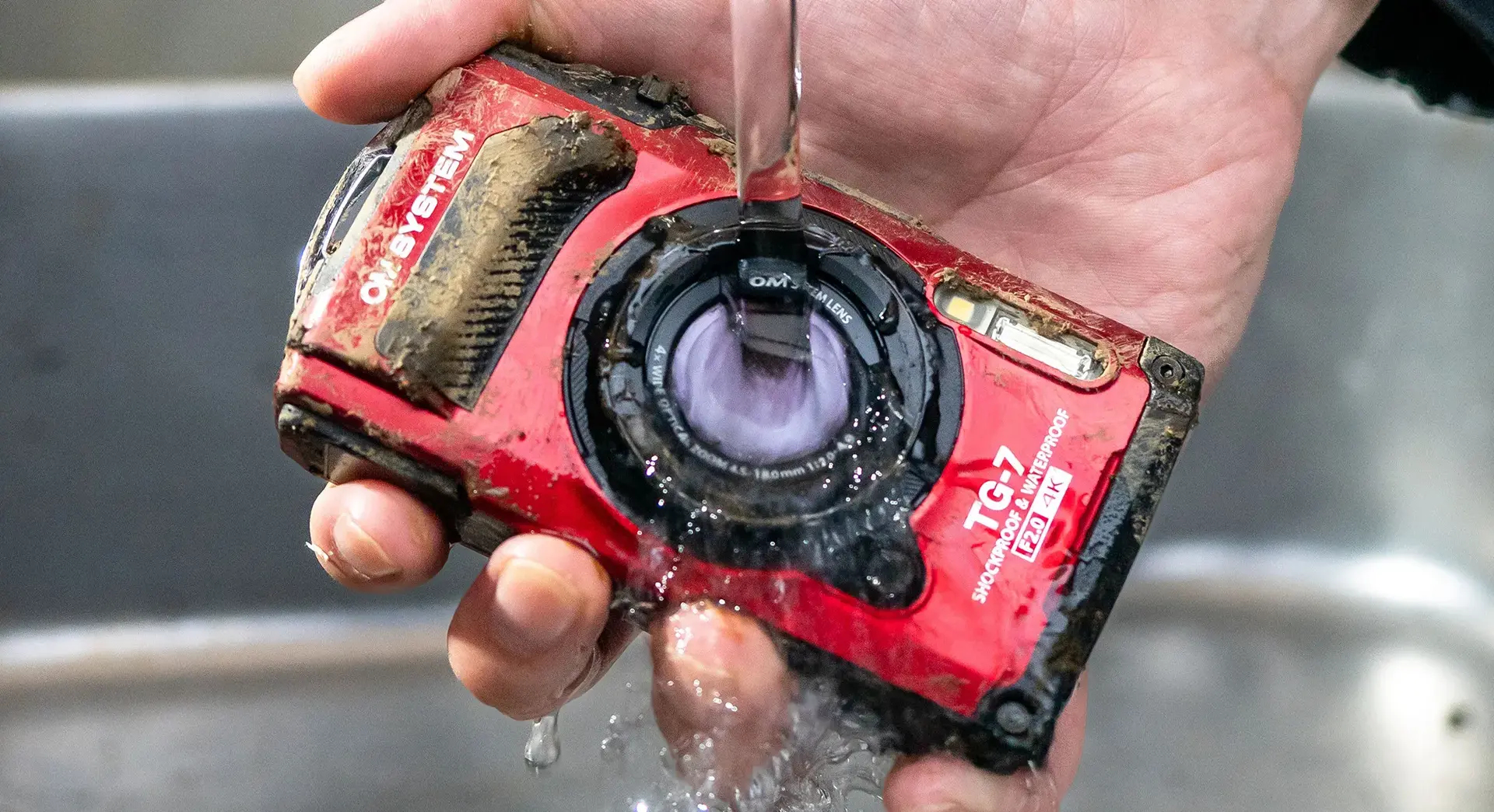 Una cámara roja OM System TG-7, cubierta de barro, se enjuaga bajo agua corriente.