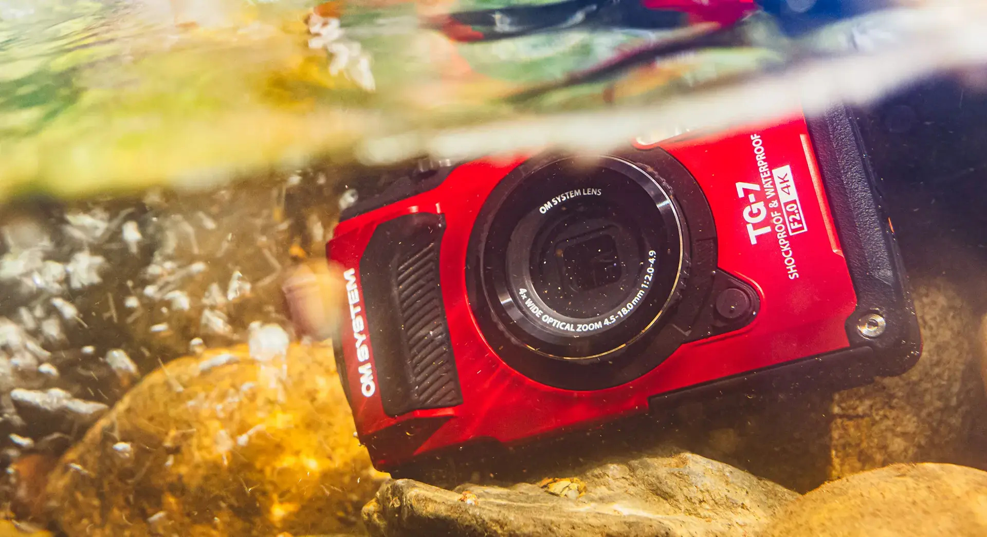 Una cámara roja OM System TG-7 bajo el agua, apoyada en rocas. Burbujas flotan alrededor.