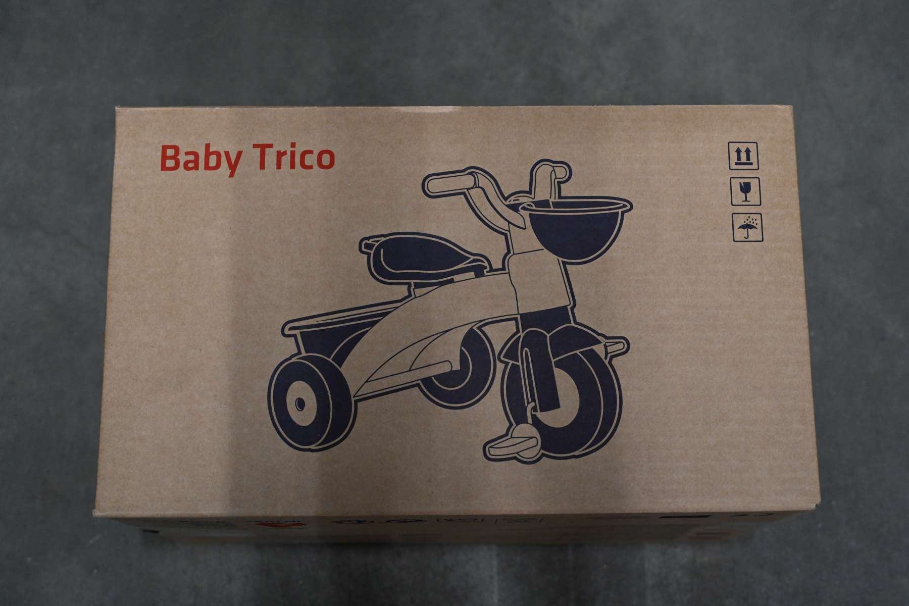 Karton mit blauer Baby-Dreirad-Illustration. Text 'Baby Trico' darüber. Vorsicht Symbole.