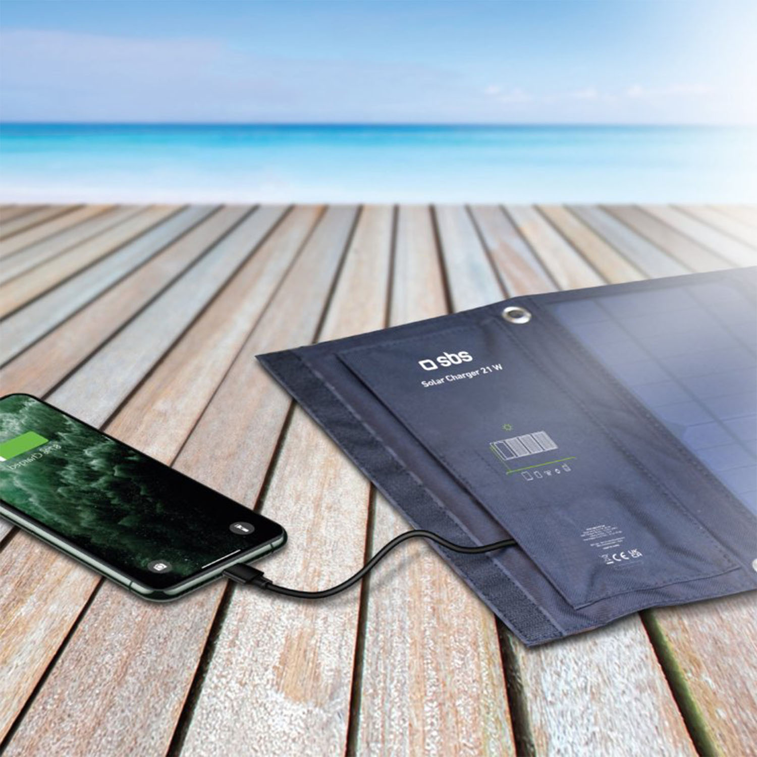 Uno smartphone si carica tramite un pannello solare su una terrazza di legno con spiaggia e oceano sullo sfondo.