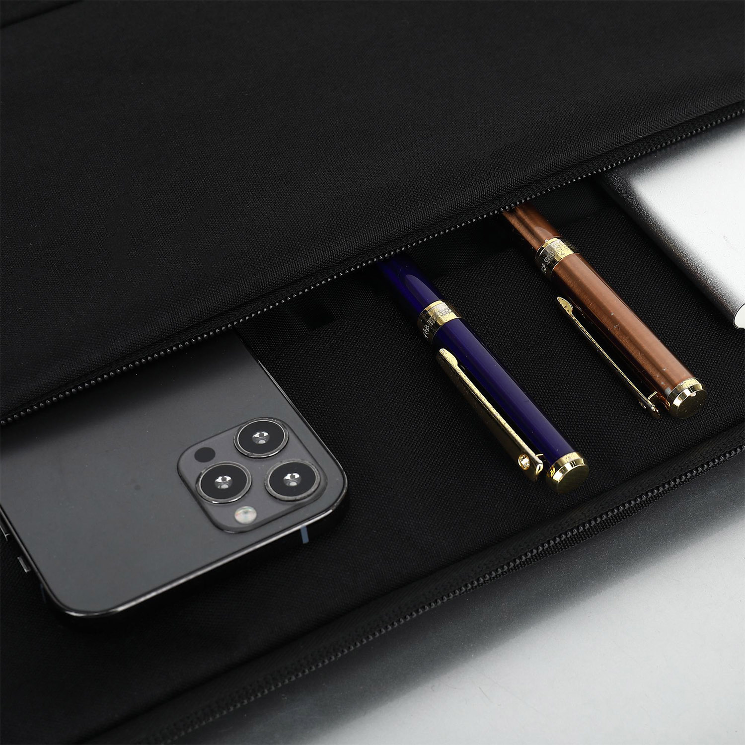 Interno borsa nera con smartphone, penne blu e marroni, e dispositivo argentato.