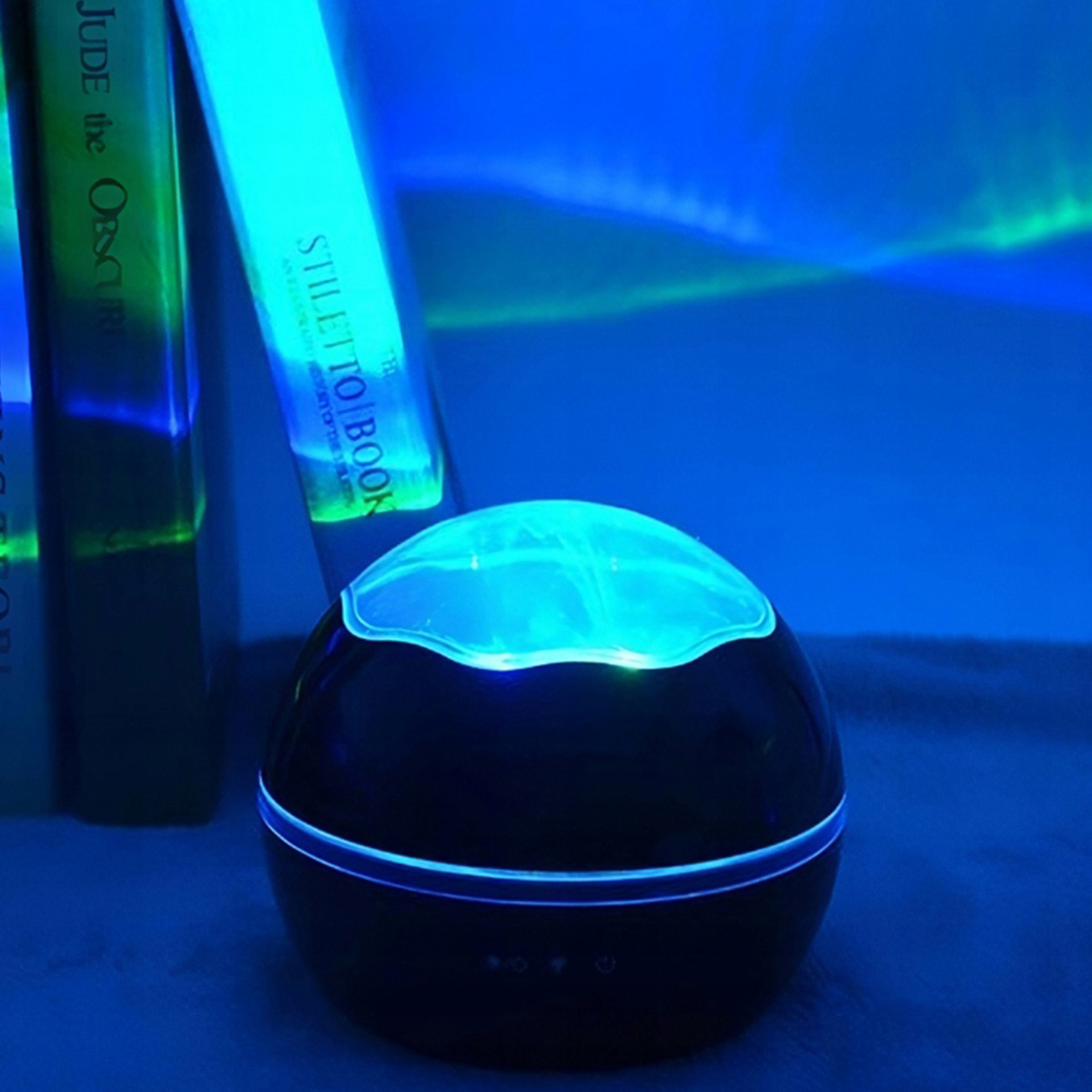 Ein schwarzer Projektor leuchtet blau und wirft aurora-ähnliche Lichter auf eine Wand nahe Bücher.