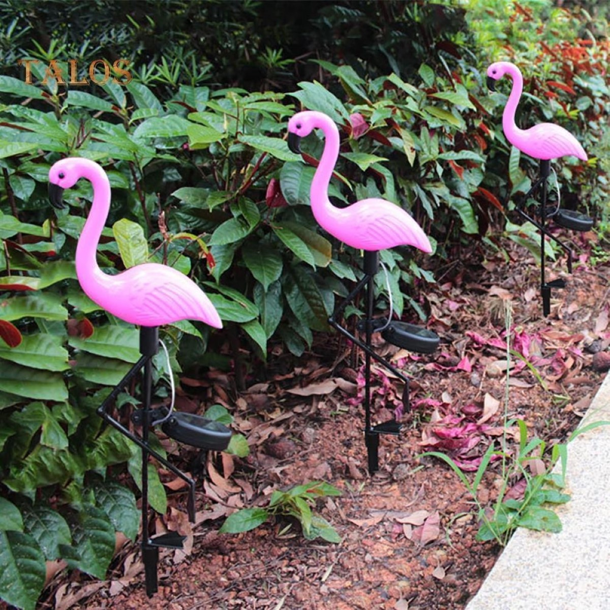 Tres luces solares de flamenco rosa se alzan en un jardín con vegetación y tierra.