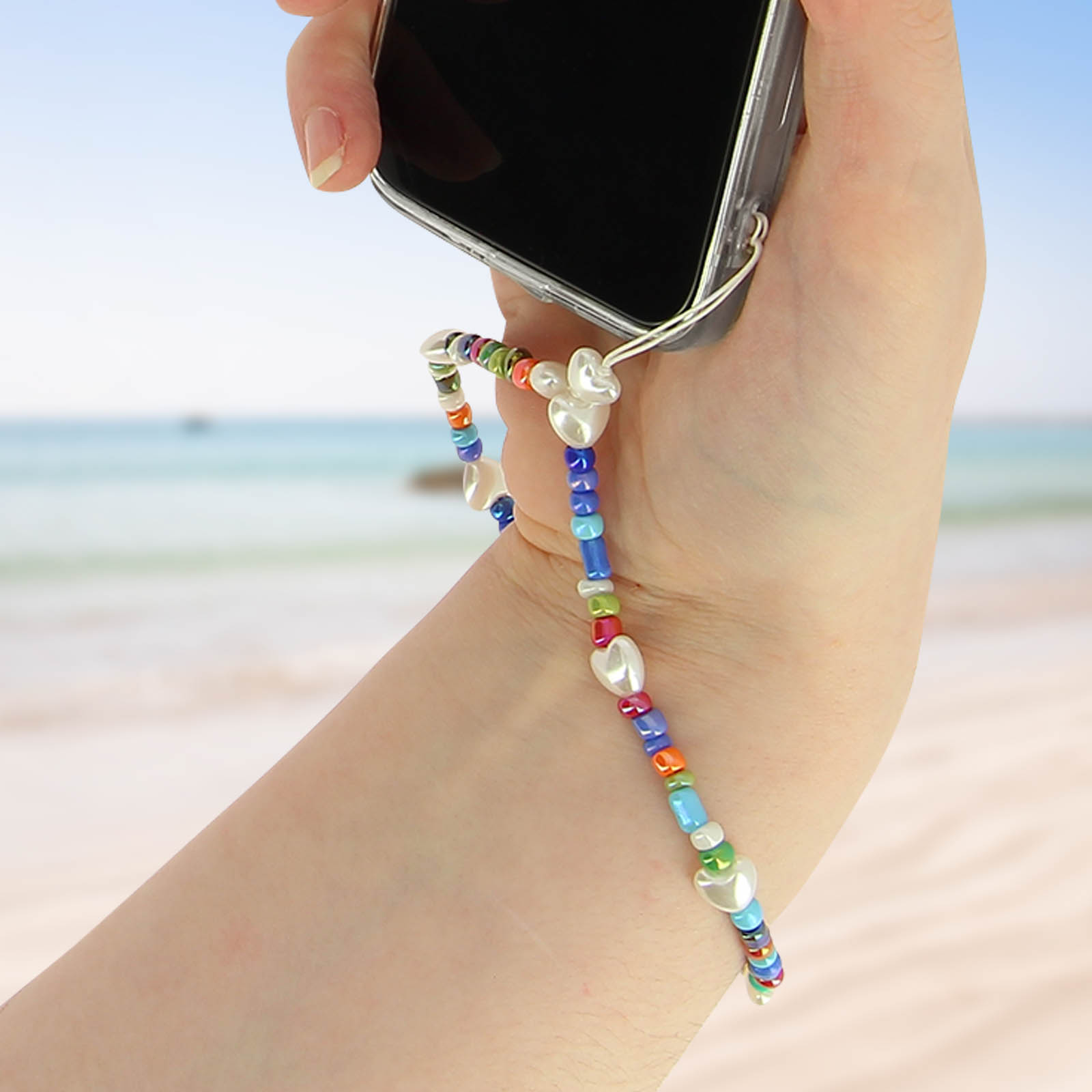 Una mano tiene un telefono con un cinturino di perline colorate con ciondoli a forma di cuore, sfondo spiaggia.