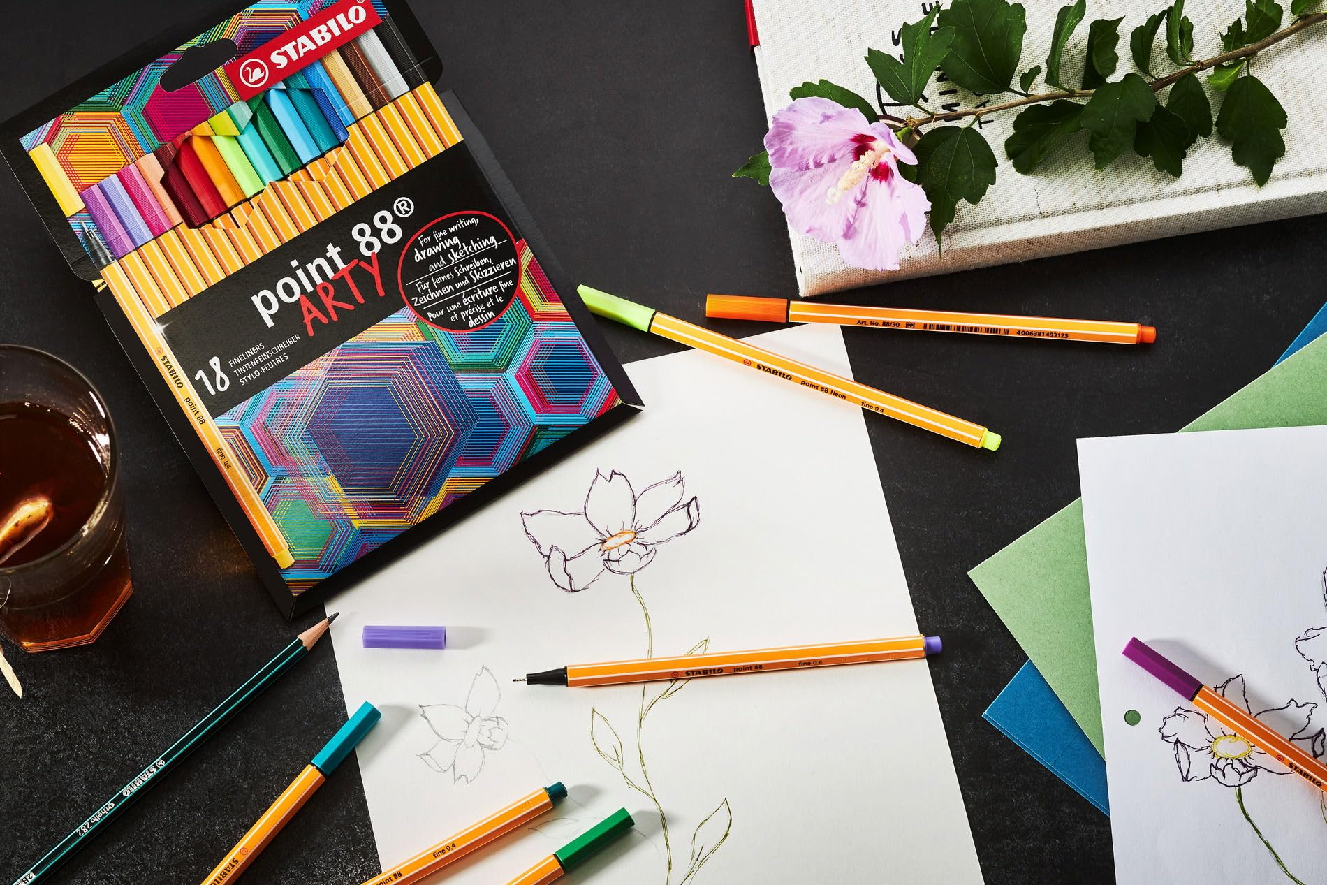 Set di penne con penne colorate, schizzi, libro e fiore su sfondo scuro.