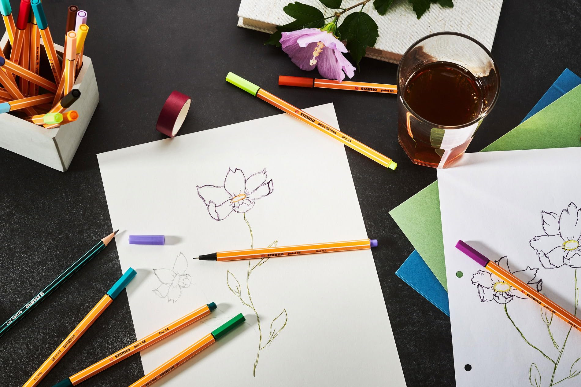 Materiali artistici: penne, matite, carta con schizzi di fiori, nastro, bevanda.