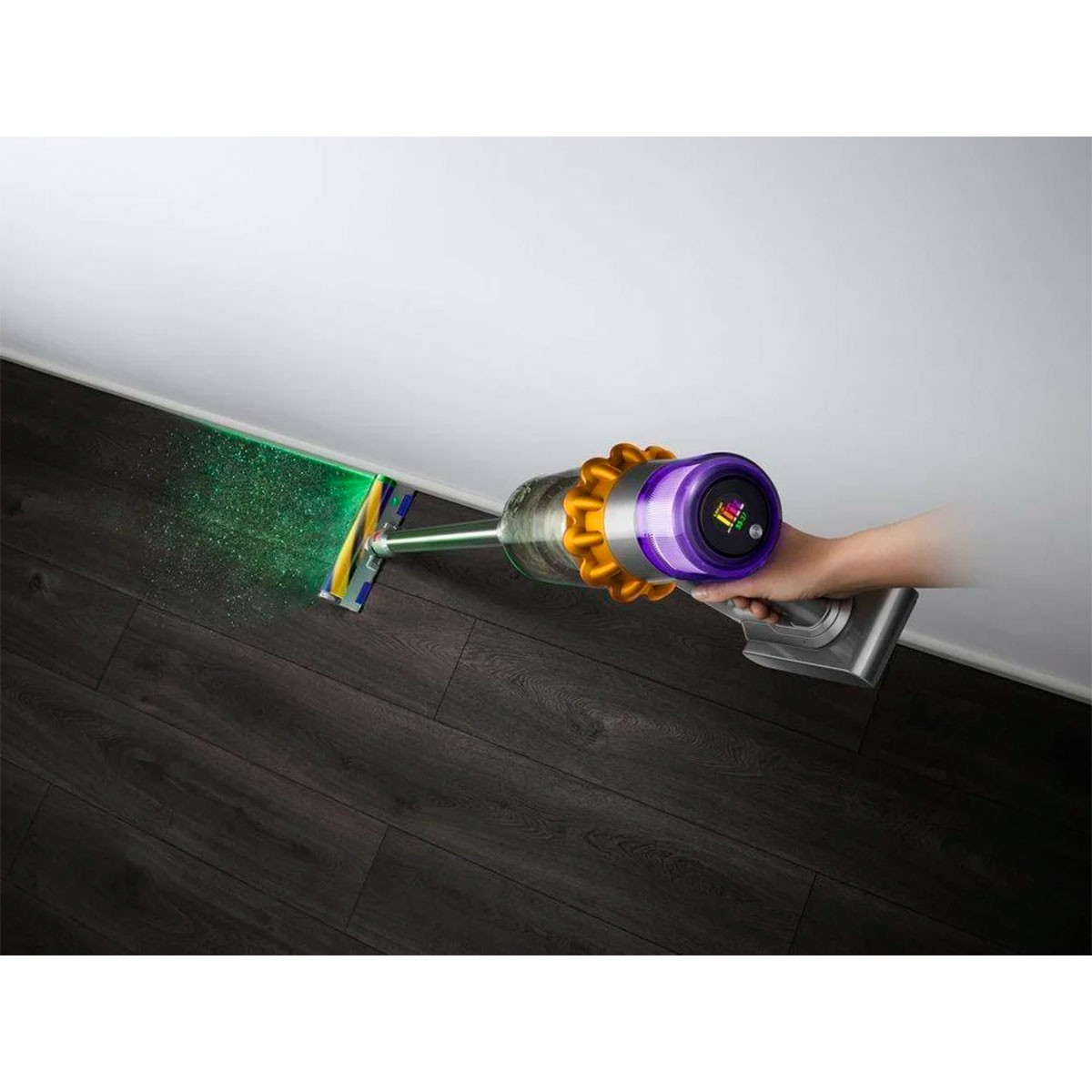 Una mano tiene un aspirapolvere Dyson viola e giallo, che pulisce un pavimento in legno scuro con luce verde.