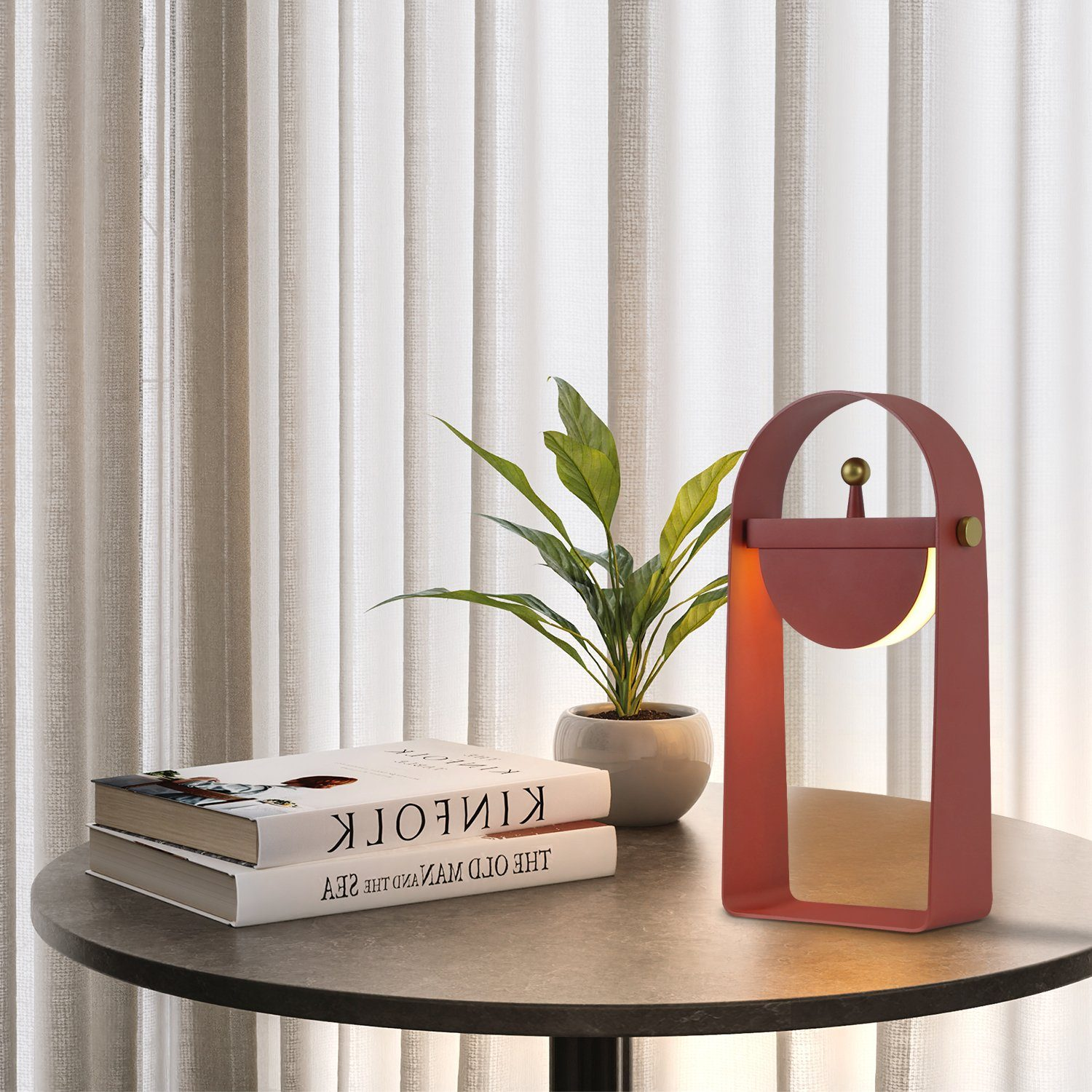 Una lampada moderna rossa, due libri e una pianta in vaso su un tavolo rotondo.